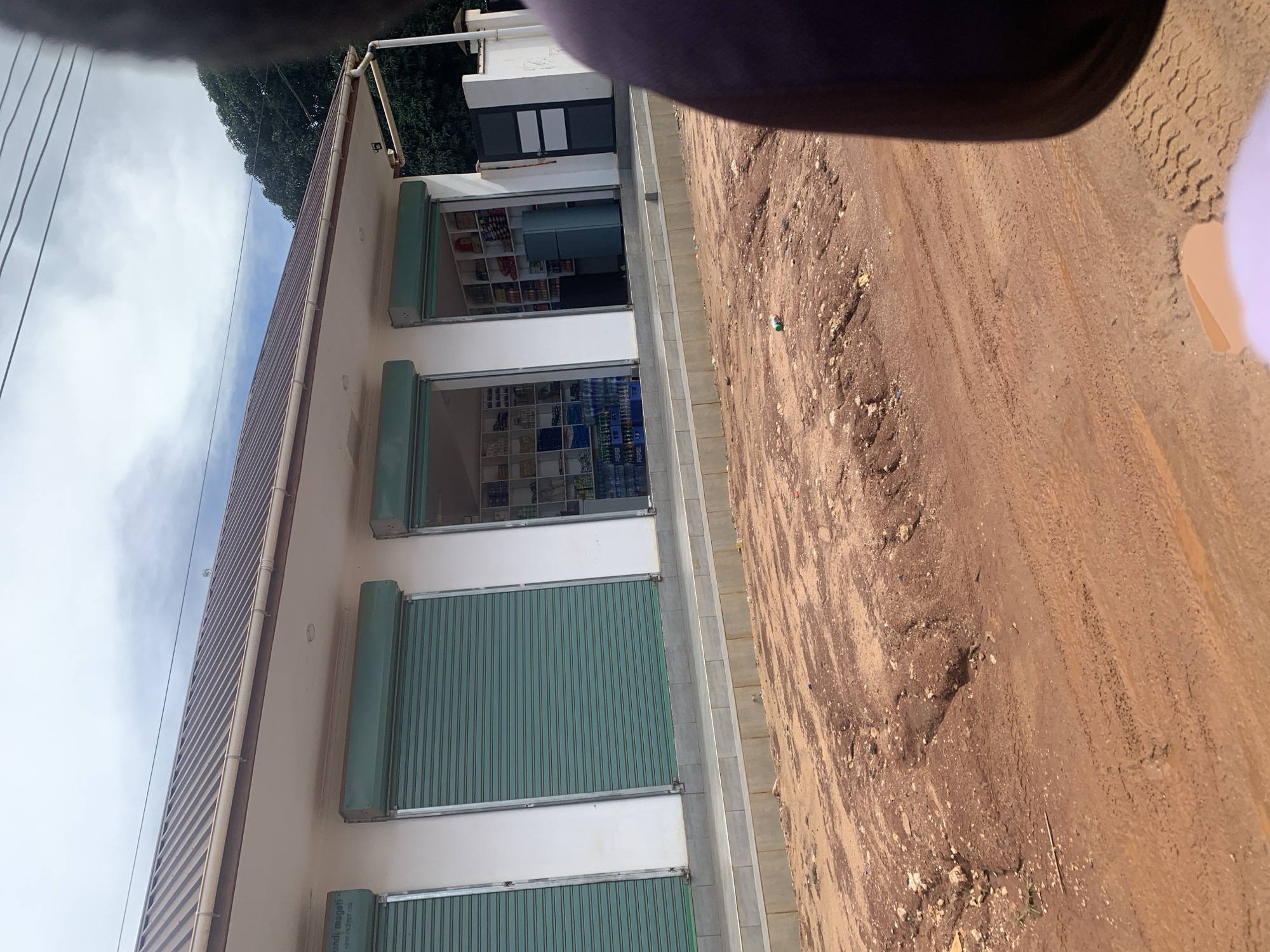 This image depicts a street scene in Dar es-Salaam, Tanzania, focusing on a single-story commercial building and an unpaved road. The building features a brown corrugated metal roof and white facade, with three distinct storefronts. The two leftmost storefronts have teal-colored rolling security shutters fully closed. The two rightmost storefronts have their shutters open, revealing retail interiors. The shop second from the left displays shelves stocked with various packaged goods and bottled beverages, including rows of Pepsi bottles, and a blue-grey refrigerator. The shop on the far right also shows shelves with diverse packaged items.
A raised concrete sidewalk or platform runs in front of the shops. To the far right of the building, a dark grey metal gate is visible, part of a perimeter wall, with dense green foliage behind it. Overhead, power lines traverse the scene against a cloudy sky.
The foreground is dominated by a reddish-brown dirt road, heavily rutted and uneven, with visible tire tracks. A small green bottle lies on the ground in the middle distance. Puddles of muddy water are present in the immediate foreground, reflecting tire treads. A partial view of a person's shoulder or the interior of a vehicle, clad in a purple garment, occupies the far right side of the frame.