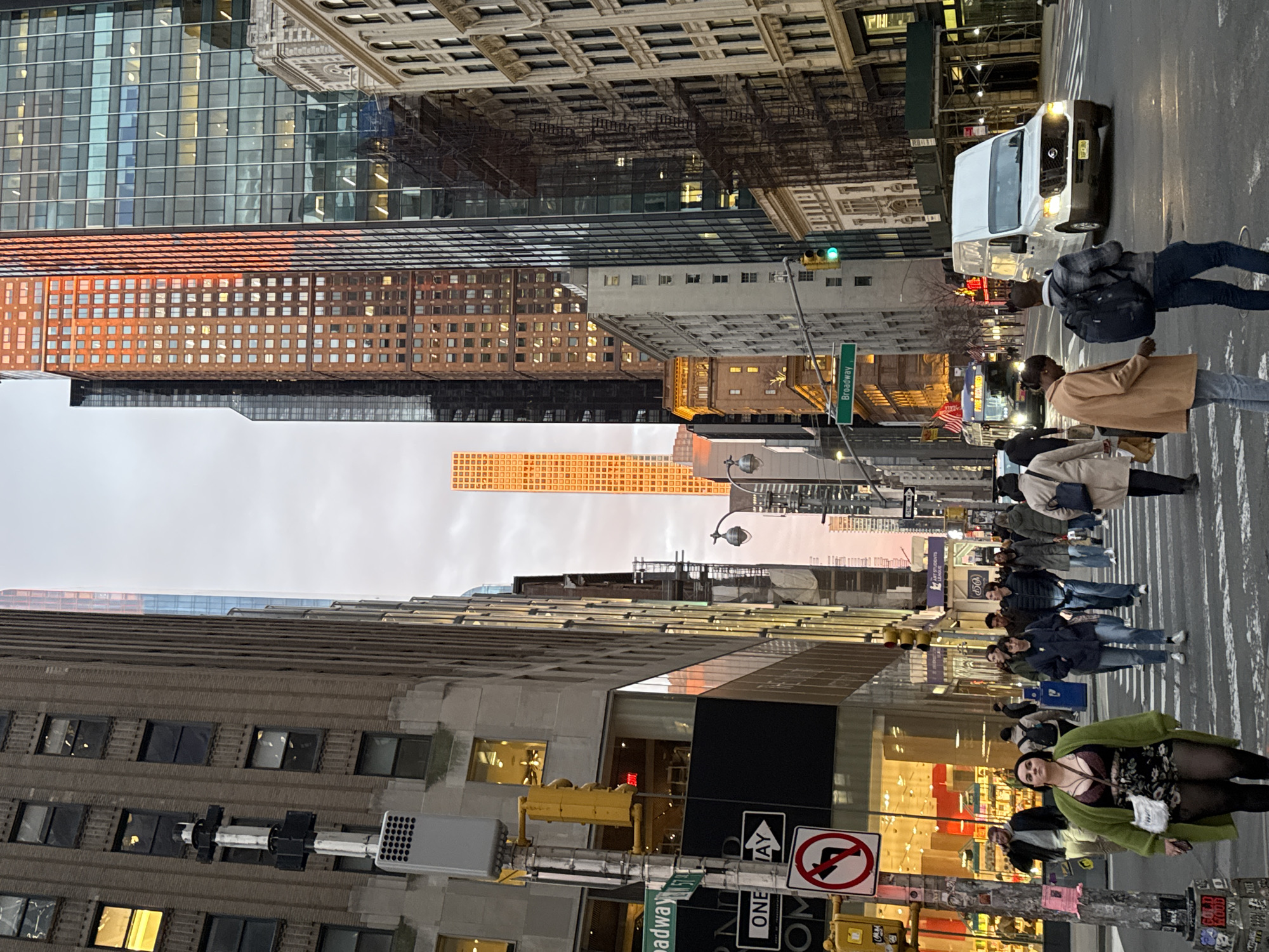 An urban street scene is depicted at the intersection of Broadway and West 57th Street in New York City, United States of America, looking east along West 57th Street. The time is either dusk or dawn, with a low sun casting a prominent orange-red reflection on several tall skyscrapers, notably 432 Park Avenue in the background and 111 West 57th Street on the right, contrasting with an overcast sky.
In the foreground, numerous pedestrians are actively crossing the street on a marked white crosswalk. A woman on the left, facing slightly forward, wears a green cardigan over a dark floral top with a maroon bralette, and carries a white shoulder bag and a phone. Other pedestrians include a man with a dark backpack and a woman in a tan coat, both with their backs to the camera, positioned closer to the center.
Vehicles include a white high-top cargo van, headlights illuminated, approaching the crosswalk from the right. Further down the street, a dark city bus with its headlights on is visible, its destination sign partially reading "FIRE DEPT".
The left side of the scene is dominated by a tall, multi-story building with a brown brick facade and rectangular windows. At its base, a brightly lit retail storefront with large glass windows and red interior accents is visible. A street pole at the intersection displays yellow traffic lights, a "Broadway" street sign, a "57th St" street sign (partially visible), a white "ONE WAY" arrow sign pointing right, and a red "NO LEFT TURN" sign.
The right side of the street features a mix of architectural styles, including an older, light-colored building with ornate details, partially covered by scaffolding, alongside modern glass and brick skyscrapers. A green traffic light is visible further down the street on the right.