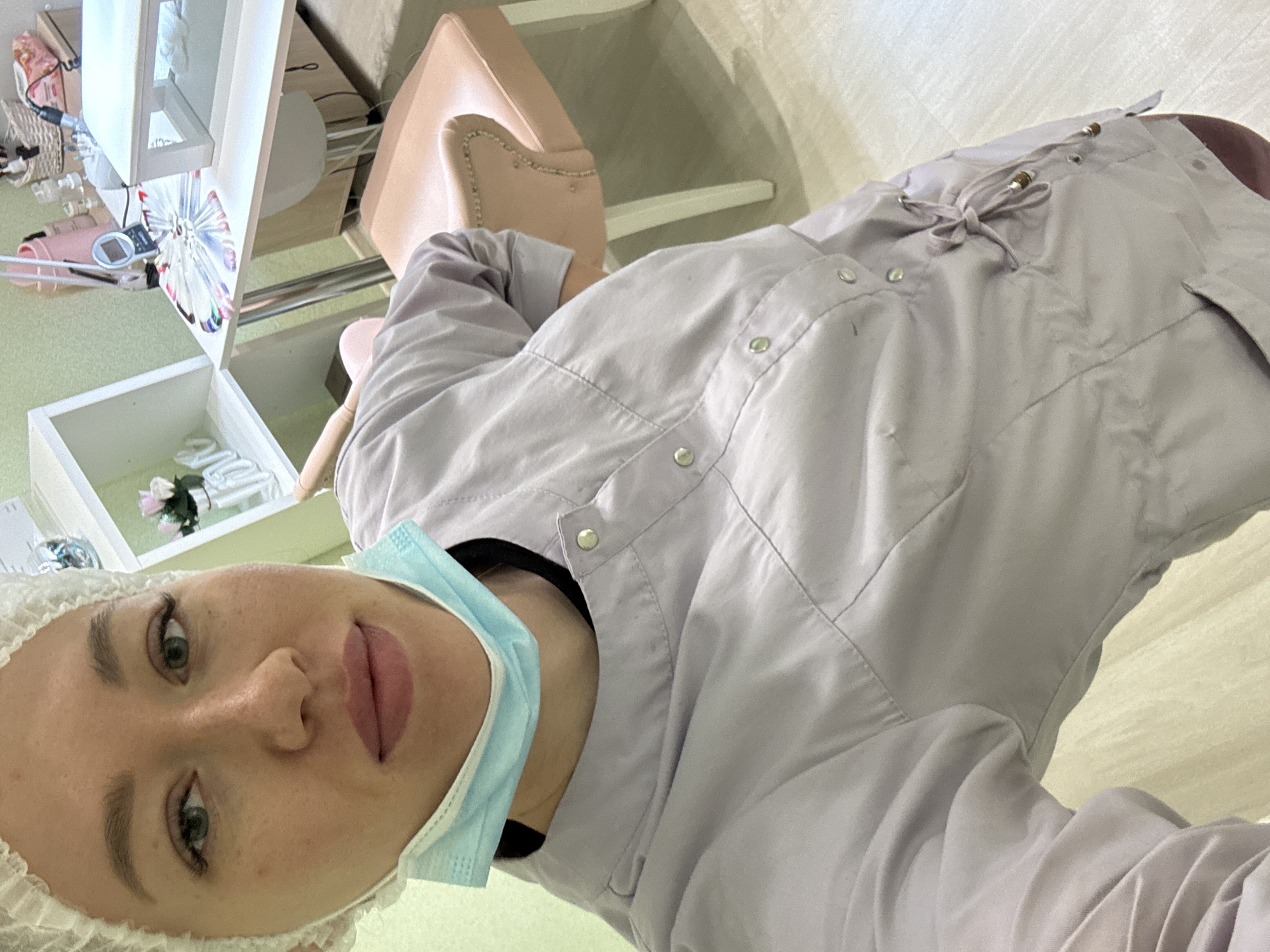 A woman wearing a surgical cap and blue surgical mask is visible, wearing a light purple button-down shirt, in what appears to be a medical or beauty treatment room. A light-colored, cushioned chair is in the background, along with a white cabinet or shelf unit. Manicuring supplies can be seen in the background. It is not possible to determine the location or city from the visual information provided.