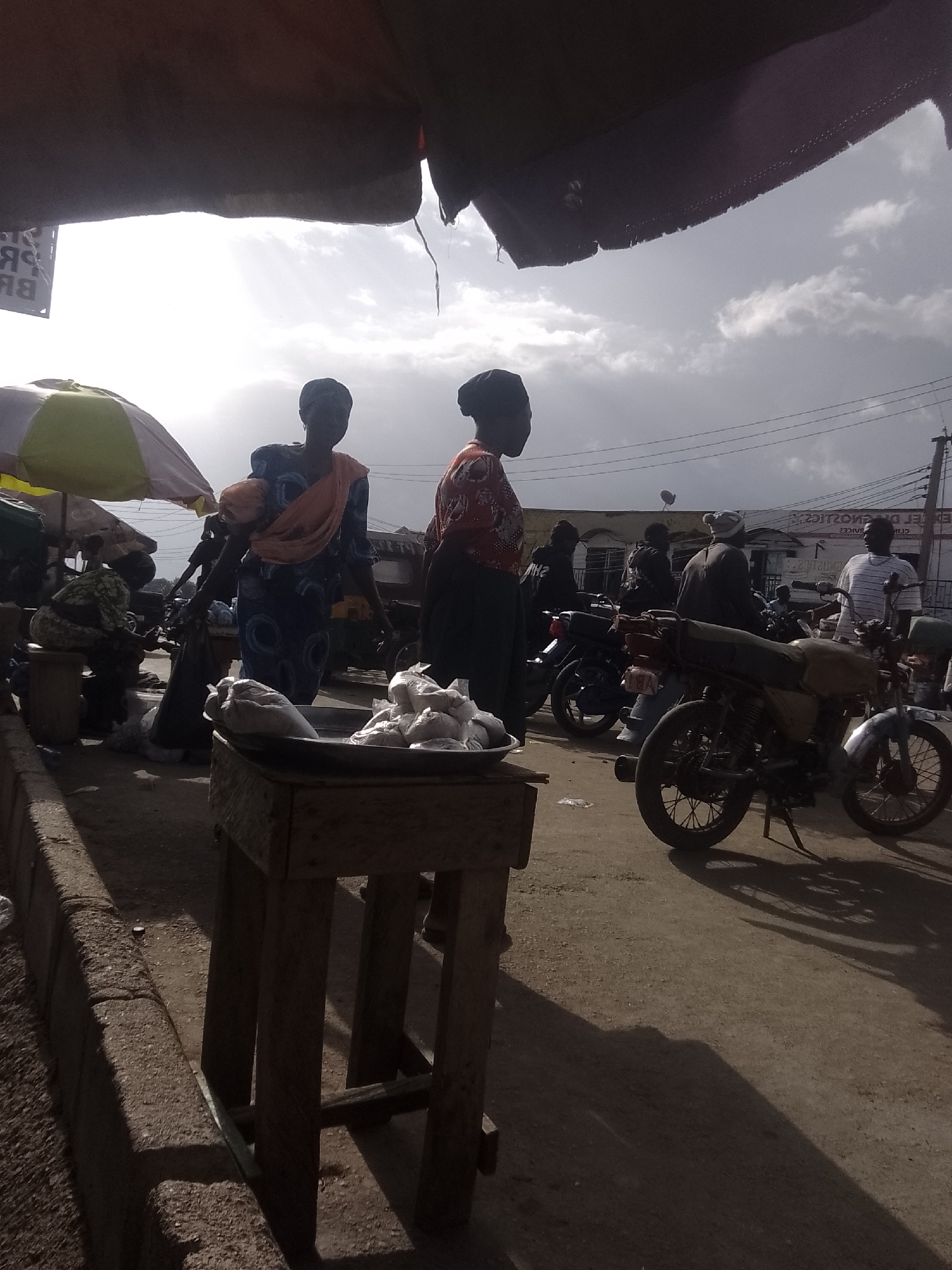 The image depicts an outdoor market scene with several people and motorcycles. In the foreground, a wooden stand holds a metal tray filled with white bags, possibly containing goods for sale. Two women are visible in the mid-ground; one in a blue patterned dress with an orange shawl, and another in a patterned red and white top. Both are silhouetted against the bright sky. Behind them, several motorcycles are parked. One motorcycle in the mid-right is a light tan color with a visible license plate. In the background, buildings with arched windows are visible, and a sign with text is partially obscured by a black awning in the upper left corner. The sky is overcast with scattered clouds, suggesting it might be daytime, possibly late afternoon due to the long shadows cast on the ground. The overall mood is that of a busy, everyday market. The text visible on the sign in the upper left corner reads "BR" and "PR".