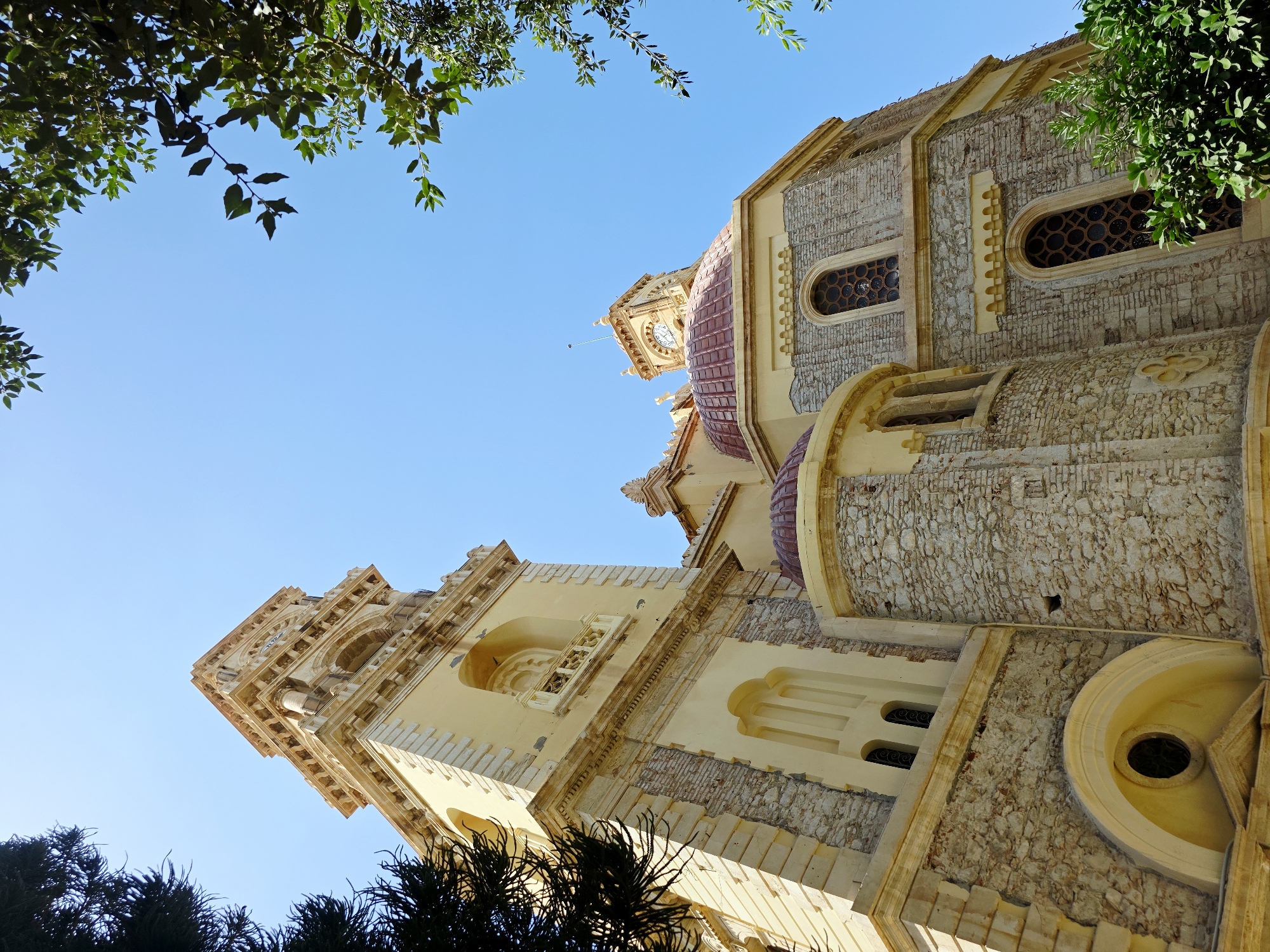 The image shows a low-angle view of the Church of Agios Titos in Heraklion, Greece. The sky is clear blue. The church is constructed of light-colored stone with cream-colored stucco accents. A tall bell tower, adorned with decorative stonework and arched openings, dominates the left side of the frame. Below the bell tower, a rounded arch frames a small circular window. To the right, a section of the church features a red-tiled dome and a clock tower. Arched windows with ornate grills are visible on the stone facade. Green foliage from trees partially obscures the top and right edges of the image. The architecture suggests historical significance and a Mediterranean setting.