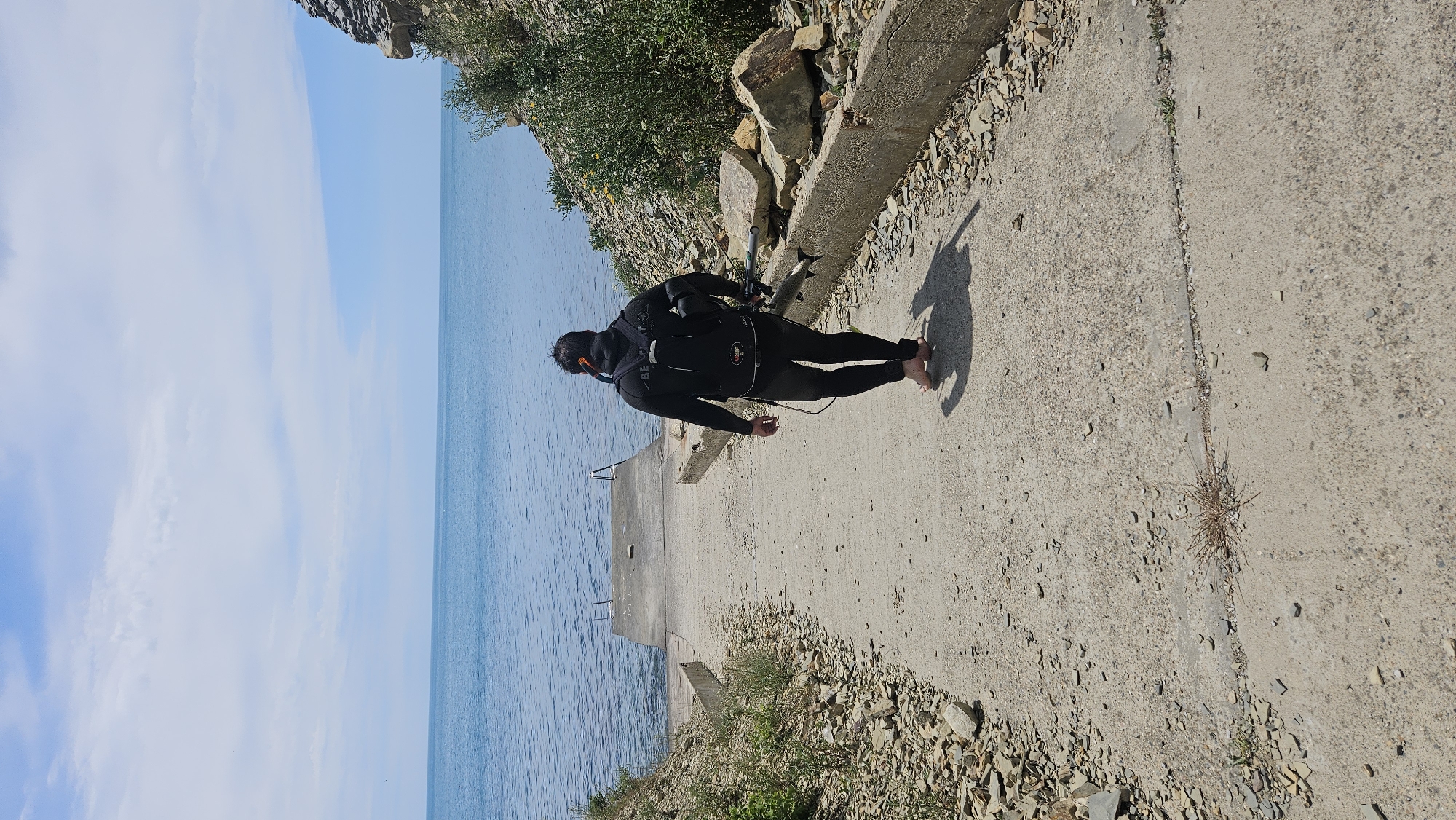 The image depicts a concrete ramp leading down to the sea. A person, dressed in a black wetsuit and backpack, is walking barefoot down the ramp towards the water. They are carrying spearfishing equipment. The sea is calm and blue, with a clear sky overhead. The ramp is bordered by rocky terrain and sparse vegetation. The scene appears to be located in Dyurso, Russia, a coastal village known for its beaches and diving spots.