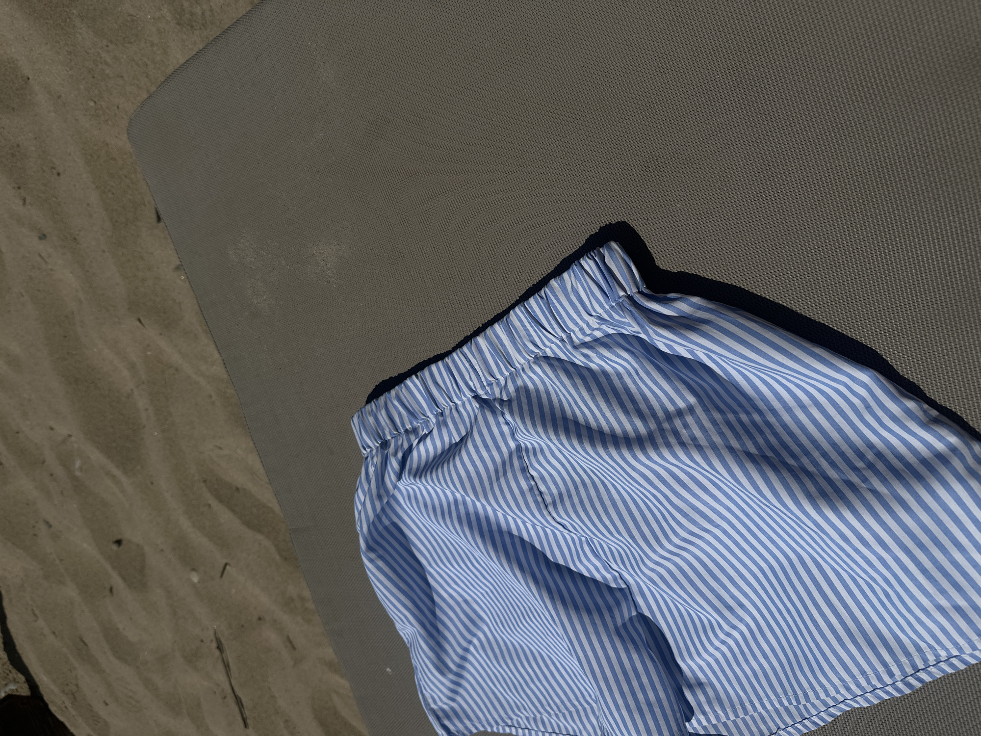 A pair of light blue and white striped shorts are laid out on a dark gray textured surface. The shorts are made of a light fabric and appear to be a casual style, possibly for lounging or beachwear. The surface they are on is likely part of a beach chair or lounger, given the sandy background. The sand behind the lounger is light brown and shows ripples and textures indicative of a beach environment. The lighting suggests a sunny day, casting shadows from the shorts onto the gray surface. The overall scene depicts a relaxed, outdoor setting, likely Ofrynio Beach, Greece.