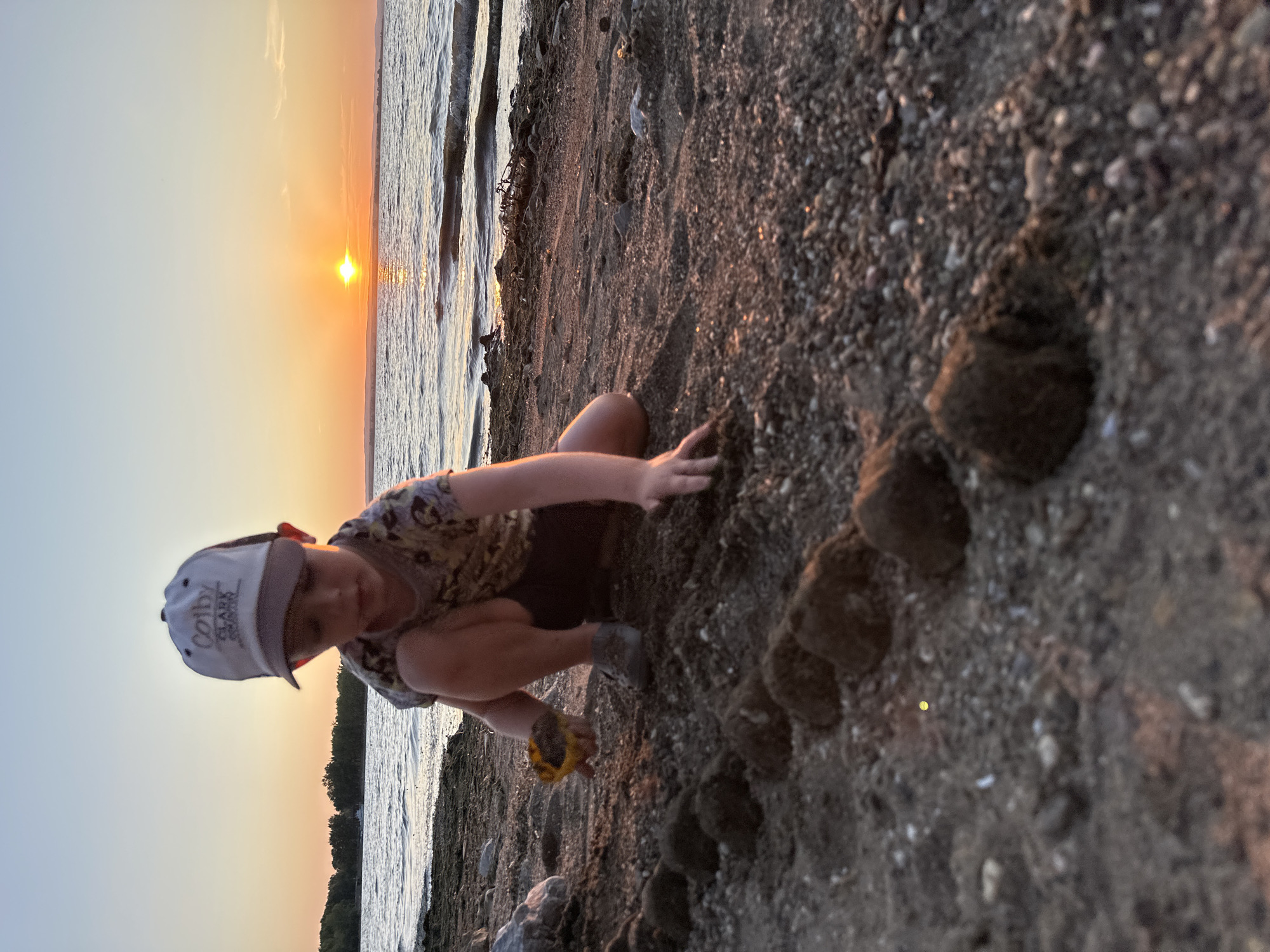 An outdoor, low-angle, medium shot captures a young child playing on a beach at sunset in Energetik, Russia. The child, wearing a cap and a patterned shirt, is crouched on the sand, focusing on a line of dirt balls they are creating. A yellow toy shovel rests near their left hand. The background features calm water with gentle waves, a distant tree line, and the sun setting over the horizon, casting an orange glow across the sky and water. The beach itself is composed of sand and small pebbles.