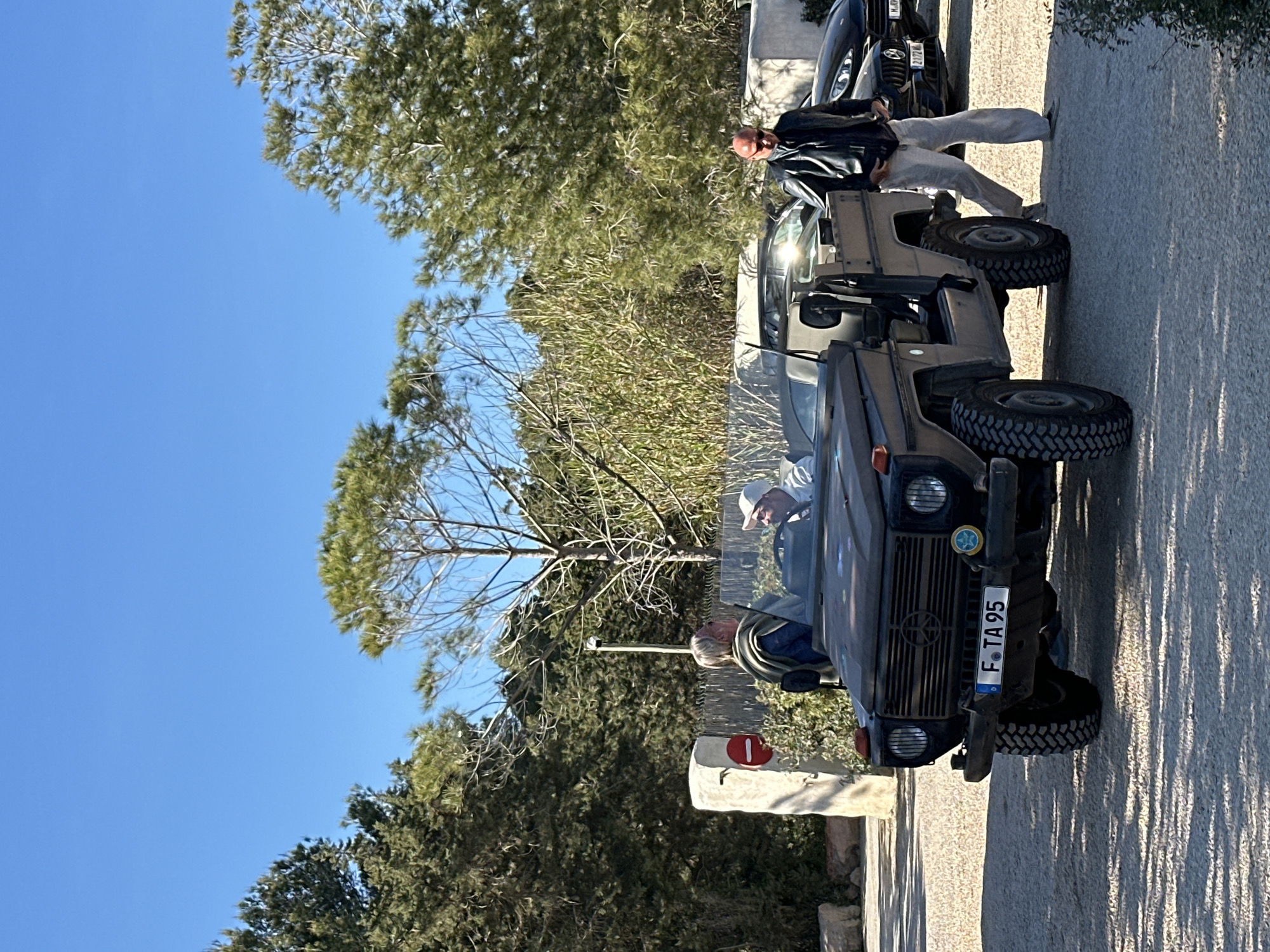 The image shows a dark-colored Mercedes-Benz G-Class parked on a gravel road with a driver and two passengers in the Sant Josep de sa Talaia. The license plate reads "F-TA 95." The backdrop consists of trees, bushes, and a clear blue sky. A "Do Not Enter" sign is visible near the car's front bumper. A man in a black jacket and light pants stands near the rear of the vehicle. Another parked car is partially visible in the background. The scene is captured in broad daylight, casting shadows on the ground.