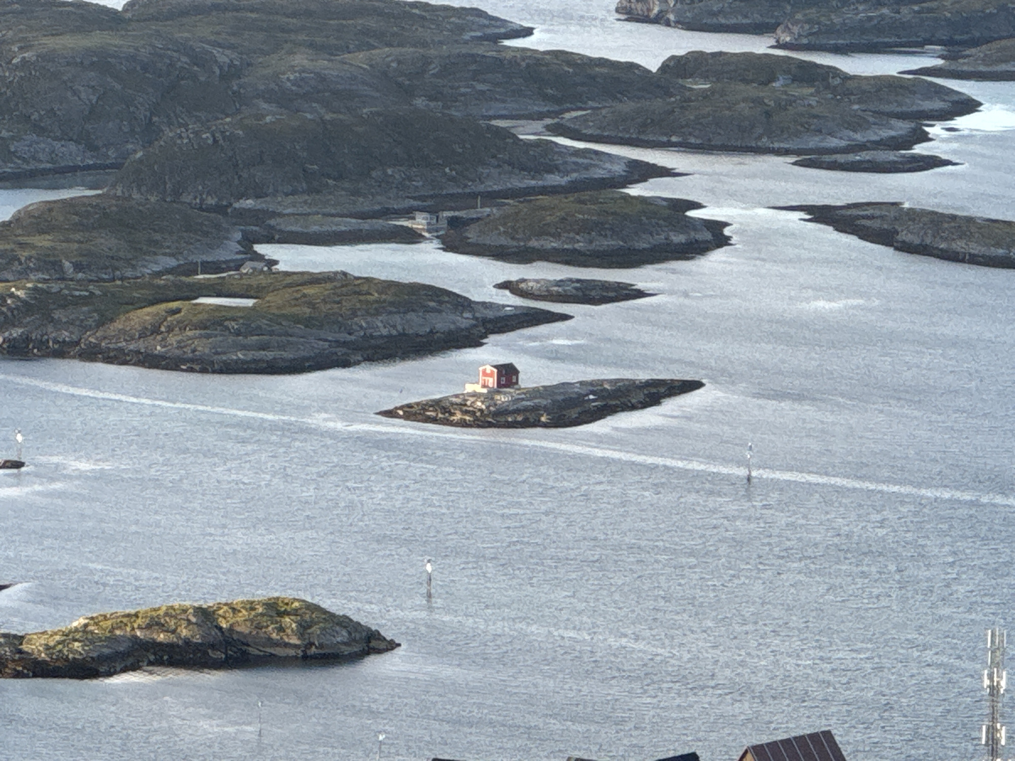 The image shows a view of the coastal landscape of Lovund, Norway, characterized by a scattering of small, rocky islands and the vast expanse of the ocean. In the foreground, to the left, a cluster of moss-covered rocks emerges from the water. Further into the scene, a prominent red building with a white trim stands on a narrow, dark, rocky islet. Several white buoys are visible in the water, marking channels or hazards. The surrounding islands are rugged and predominantly covered in dark, weathered rock, with patches of green vegetation. The water itself appears calm, reflecting the overcast sky. In the distance, larger islands with more pronounced topographical features can be observed. The overall impression is one of a remote and serene coastal environment.