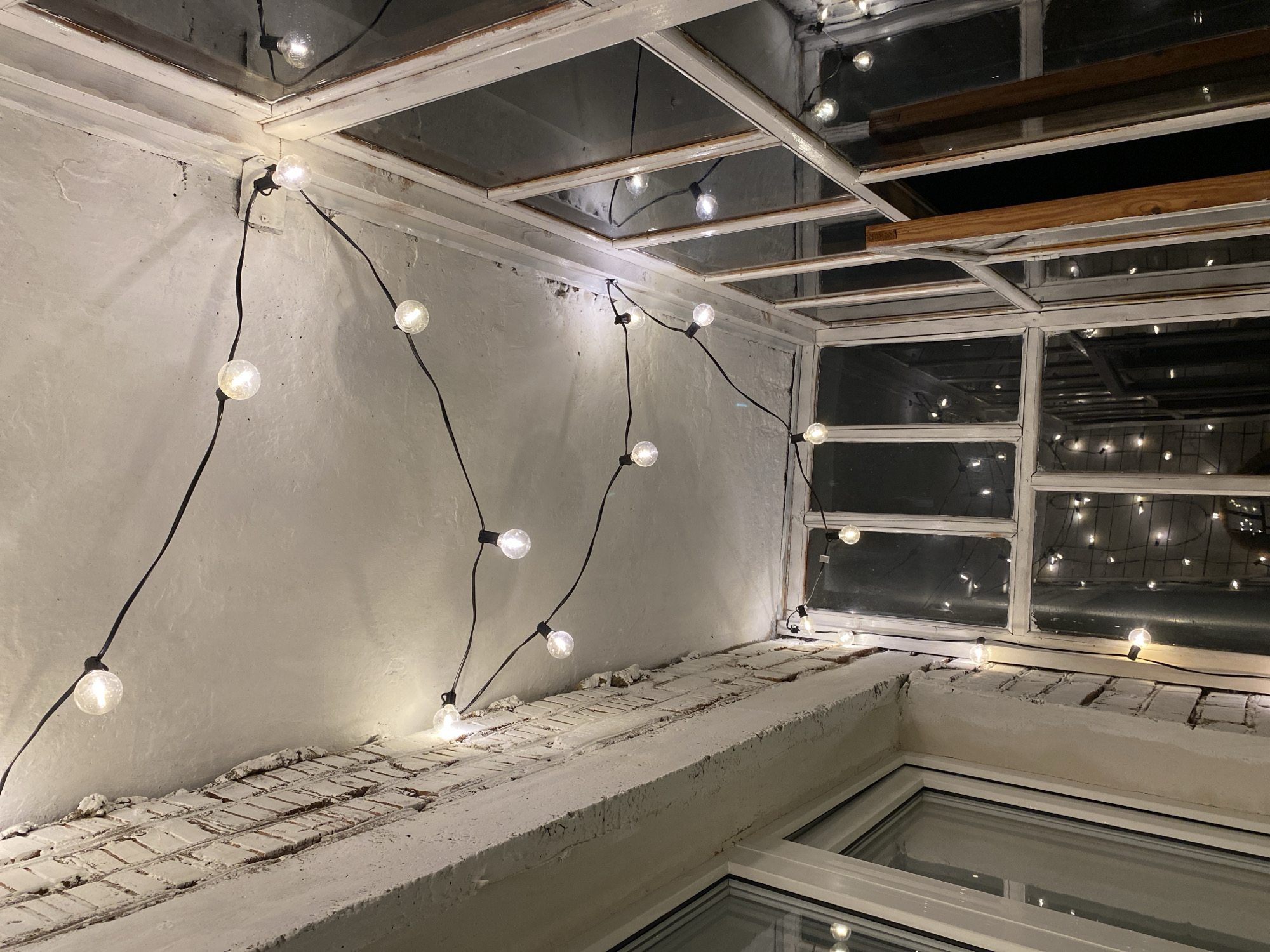 This is a forensic, matter-of-fact description of an indoor scene, likely a sunroom or enclosed balcony, in Votkinsk, Russia. The primary subject is a string of illuminated globe lights stretched across the white ceiling and down along a series of old, multi-paned windows. The ceiling appears to be rough-textured plaster with some cracking and discoloration. A section of exposed brick wall, painted white and showing age, is visible on the left. The windows are framed with white-painted wood, and some panes are reflective, showing the interior lights. One section of the window frame is open, revealing a darker interior space with more string lights visible. The overall impression is one of a slightly neglected but cozy space, illuminated by the warm glow of the string lights. There are no people or animals present in the image.