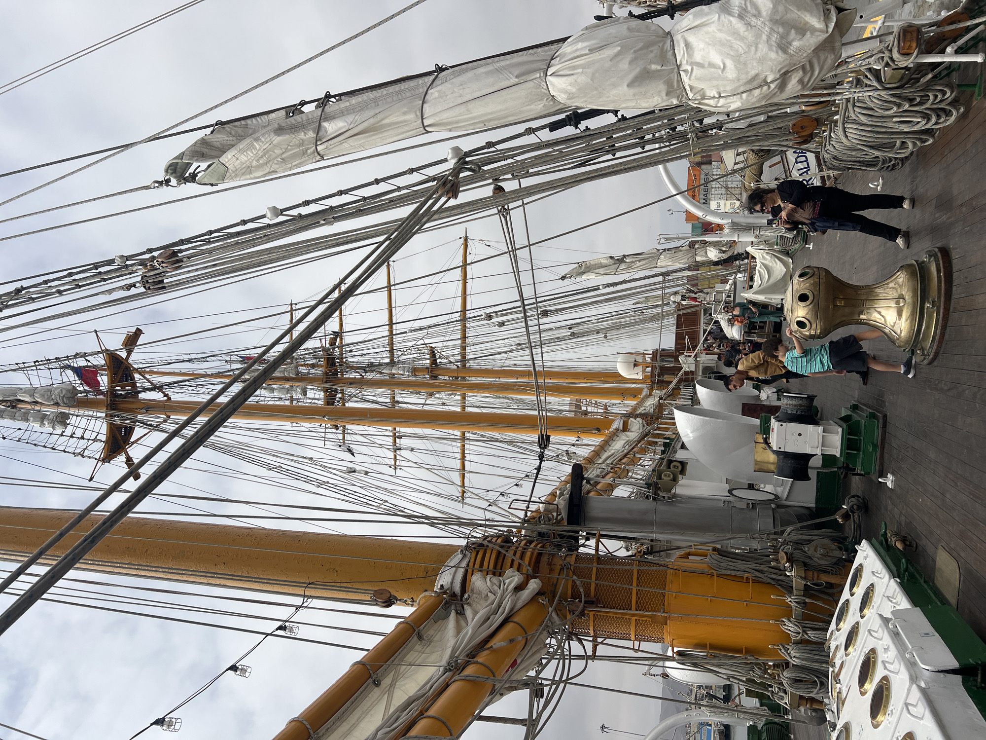 The image shows the deck of a tall ship docked in Arica, Chile. The ship's rigging and sails dominate the scene. Several people, including a child and an adult taking pictures, are on the deck. There are winches and other nautical equipment visible. The sky is overcast. The ship's hull is green and white. A cargo ship is visible in the background.