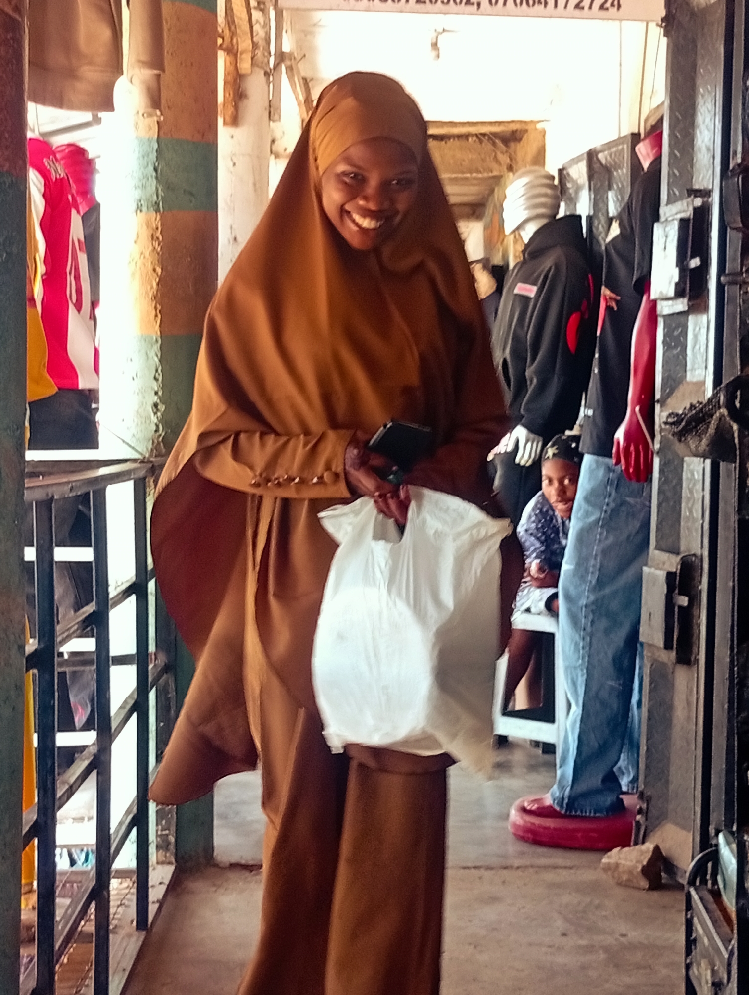 In Bukuru, Nigeria, a woman wearing a brown hijab and a long brown garment stands in a market stall. She is smiling and holding a white plastic bag in her right hand. To her left, on a rack, are several brightly colored garments, possibly clothing for sale. Behind her, a mannequin wears a black hoodie. Further back, a young person wearing a blue and white patterned shirt and jeans is partially visible. The stall has metal shelving and a railing. The ground is concrete. The overall impression is a busy market environment.