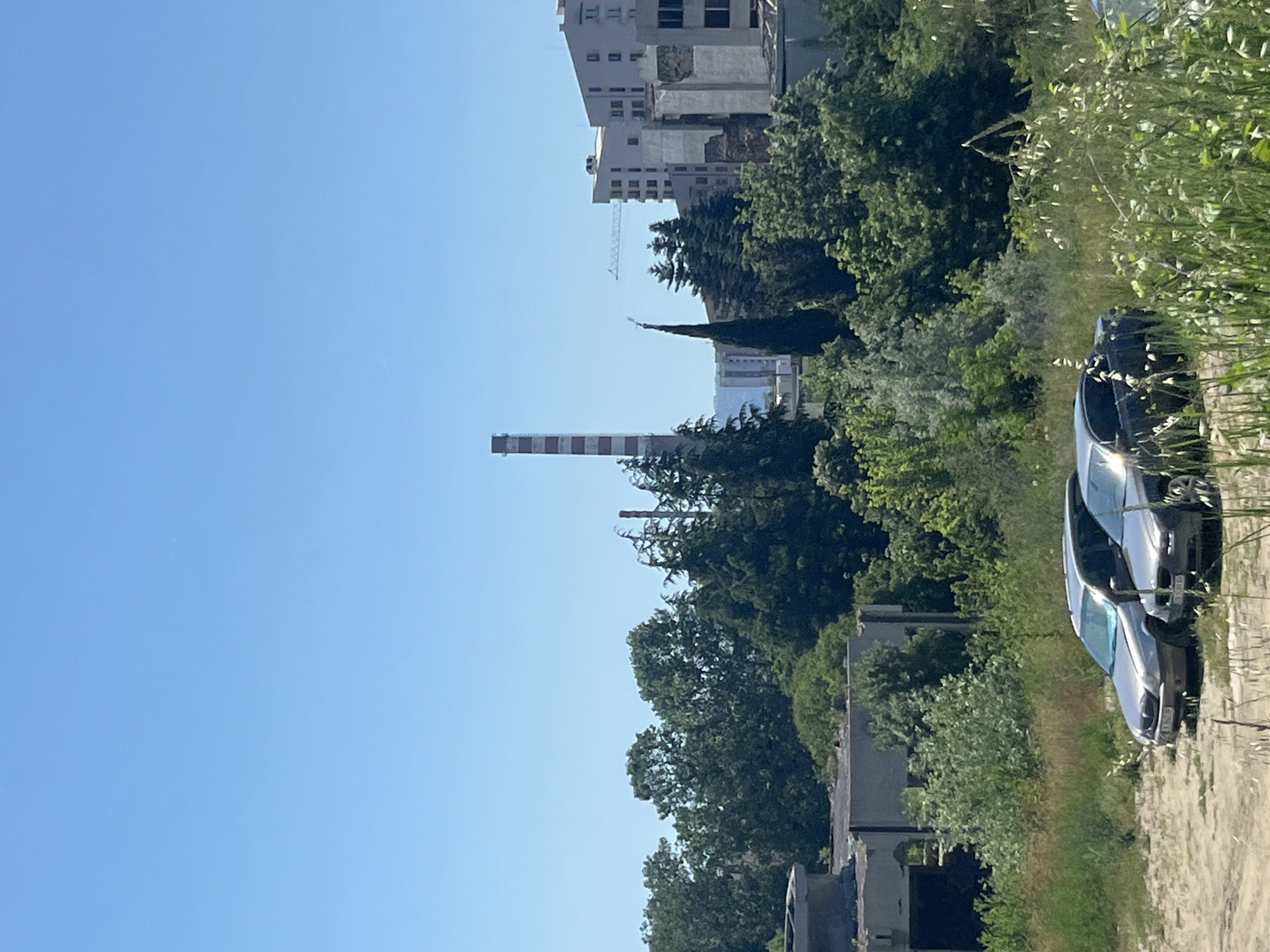 The image shows a cityscape with a clear blue sky. In the foreground, two cars are parked on a dirt lot surrounded by overgrown grass and vegetation. Behind the cars, there are several buildings, some appearing to be under construction or in a state of disrepair. Trees and foliage are abundant, creating a contrast between the natural and urban elements. A tall, red and white striped chimney or smokestack is visible in the middle ground, alongside several trees and some taller buildings. A construction crane is also present in the distance. The scene appears to be an outdoor setting, possibly on the outskirts of a city, with evidence of industrial and residential development. The vegetation suggests a temperate climate. The image does not contain any people or discernible actions.
