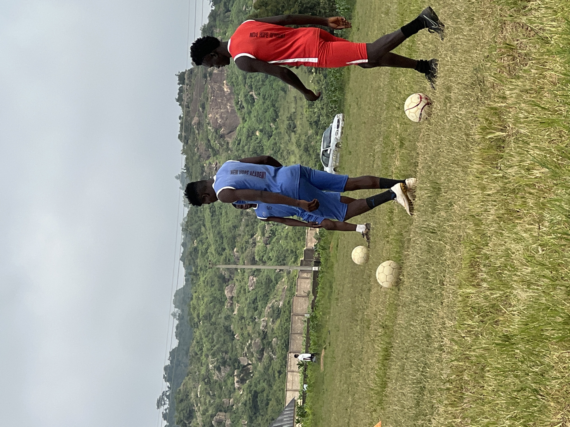 In Jos, Nigeria, on an overcast day, three young men are on a grassy field, practicing soccer. Two men, dressed in a blue and white soccer uniform and a red and white soccer uniform, respectively, are the focus of the image. The man in blue is facing away from the camera, while the man in red is walking away, his back to the viewer. A third player, partially visible, stands behind the man in blue. Several soccer balls are scattered on the field, some in play and others waiting. In the background, a white sedan is parked near a fence, and a lush green hillside dotted with rocks rises towards a cloudy sky.