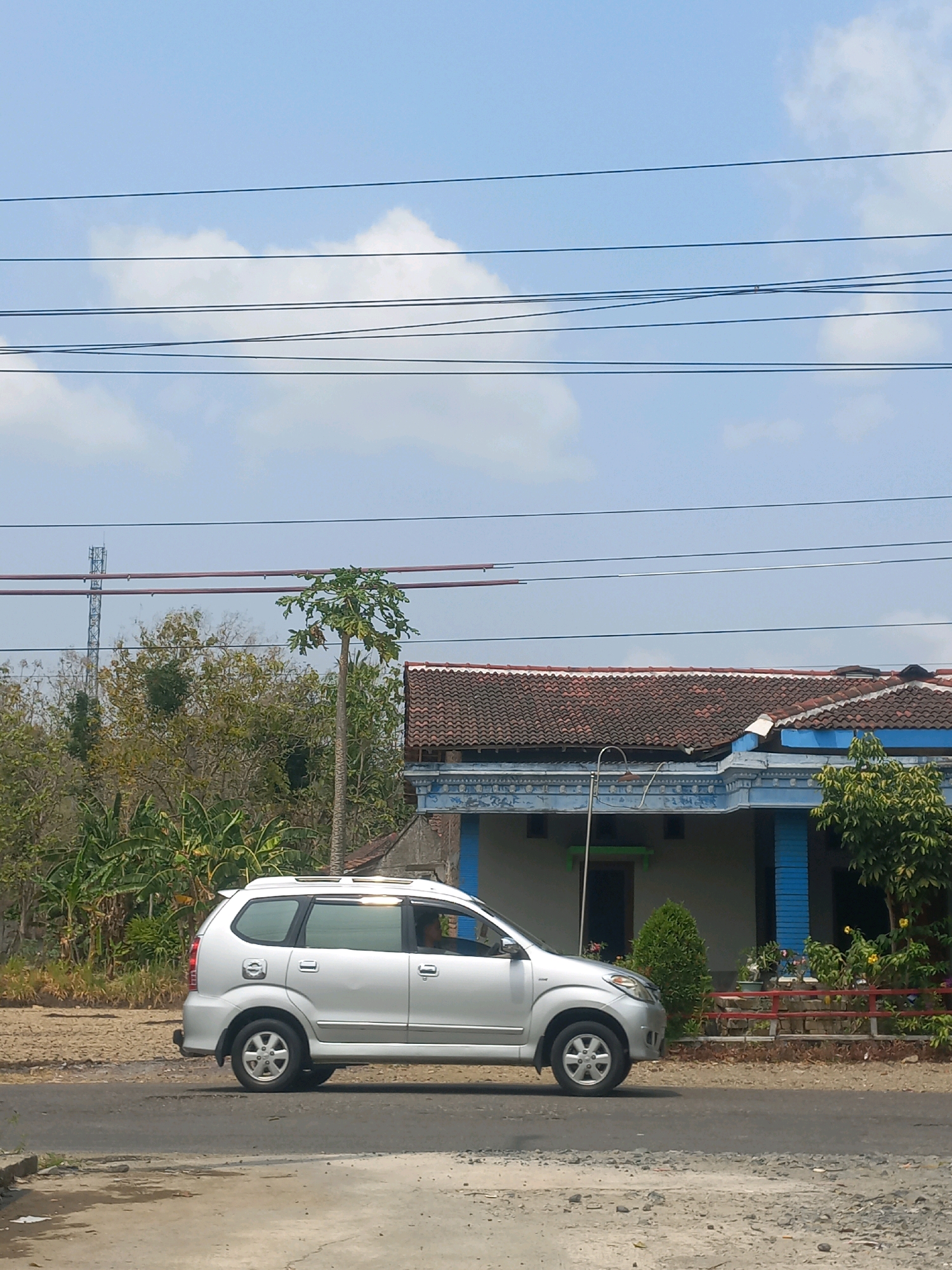 A silver minivan is driving on a paved road in a rural area with a house and vegetation in the background. The sky is blue with scattered clouds and several power lines are strung across the top of the image. The minivan is silver and has its windows tinted. A person is visible inside the minivan. To the right of the road is a single-story house with a red tiled roof and blue trim. There is a small green tree in front of the house. Vegetation, including banana trees, and a cell tower are visible behind the house. The road is cracked and appears to be made of asphalt and gravel. The scene appears to be in Baturetno, Indonesia, based on the context provided.