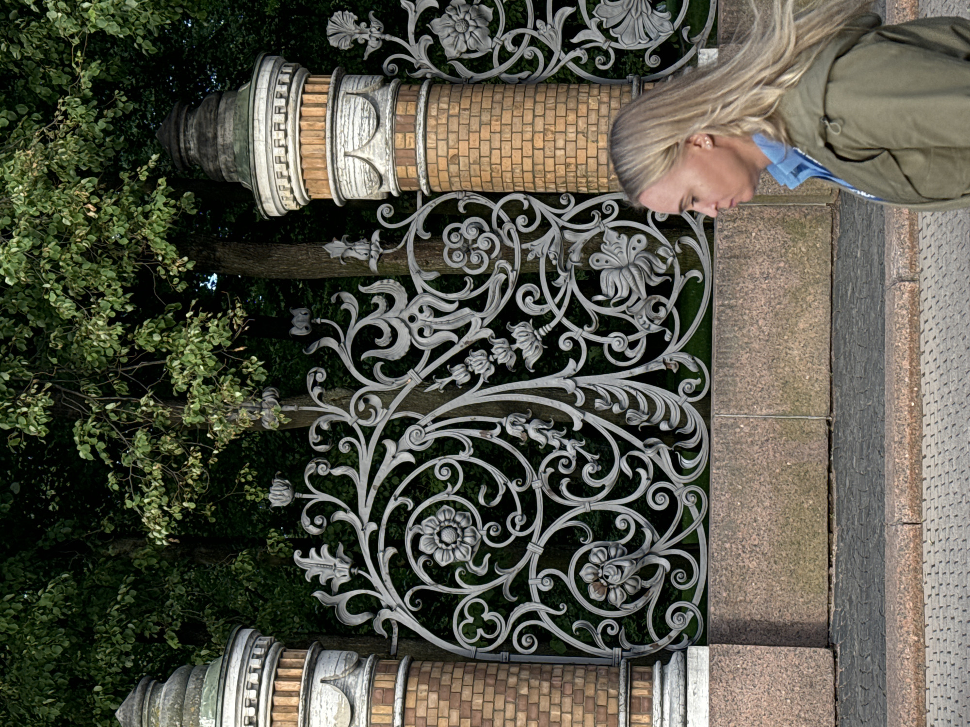 A woman with blonde hair is walking past a decorative metal fence and brick pillars. The fence has intricate floral patterns and the pillars are topped with sculpted elements. Behind the fence, there are trees with dense green foliage. The ground in front of the fence is paved with cobblestones and granite slabs. The scene appears to be an outdoor setting, possibly a park or a historic area in Saint Petersburg, Russia. The woman is wearing a green jacket over a blue shirt and is captured in profile, looking to the left.