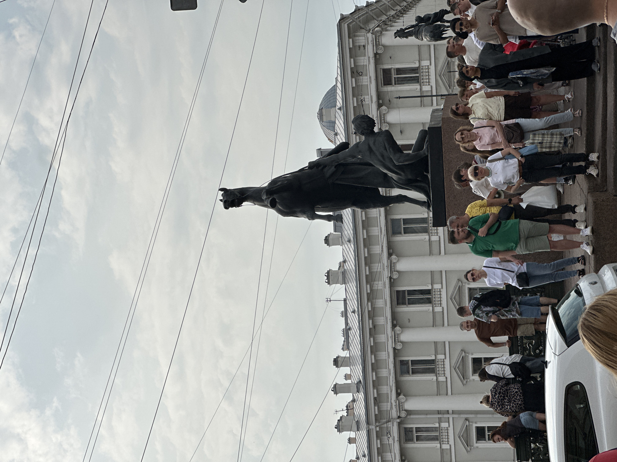 The image shows a crowd of people gathered in front of a large, dark bronze sculpture of a rearing horse and a man struggling with it. The sculpture is located on a pedestal, and in the background, a grand white building with columns and arched windows can be seen, along with a dome. This building appears to be the General Staff Building on Palace Square in Saint Petersburg, Russia. The sky is overcast with scattered clouds. The people in the foreground are dressed in casual summer clothing, and some are looking at the sculpture, while others are talking amongst themselves. A white car is partially visible in the lower left corner of the image. The overall scene suggests a tourist attraction or a public gathering space.