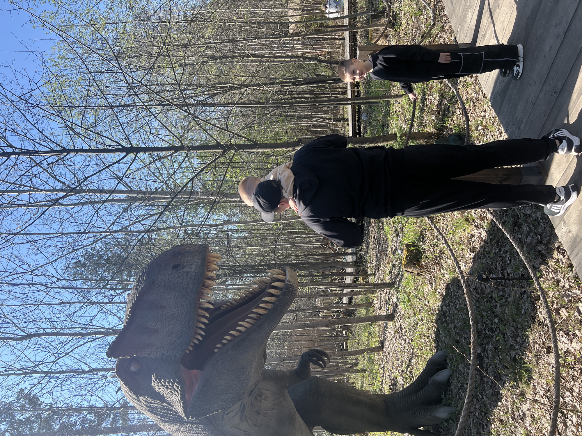 The image shows a dinosaur model with two people on a wooden walkway in a park setting. The dinosaur is a large, gray model with its mouth open, revealing teeth. A man is bent over looking at the dinosaur with a young child on his back. A second child stands on the boardwalk. Bare trees and blue sky are visible in the background. The setting is in a park in Izhevsk, Russia.