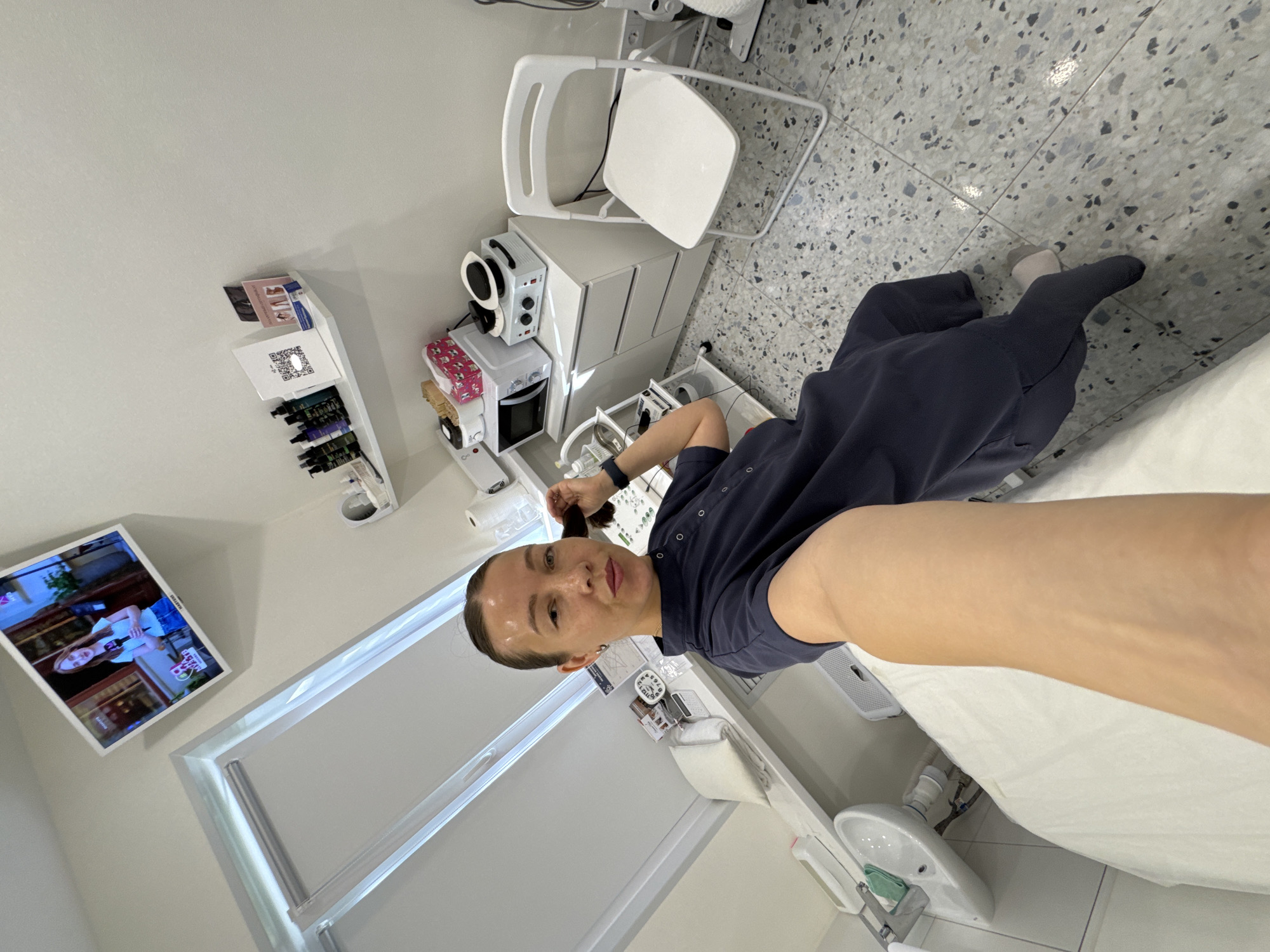 A woman is taking a selfie in a bright, clean room that appears to be a medical or beauty treatment room. The room has white walls and light-colored tiled flooring. In the background, there is a white folding chair, a cabinet with drawers, and various pieces of equipment, including what looks like a microwave, a device with knobs, and a sink. A flat-screen TV is mounted on the wall, displaying a woman speaking on screen. The woman in the foreground is wearing a dark blue uniform and is holding her hair. The overall impression is of a professional setting.