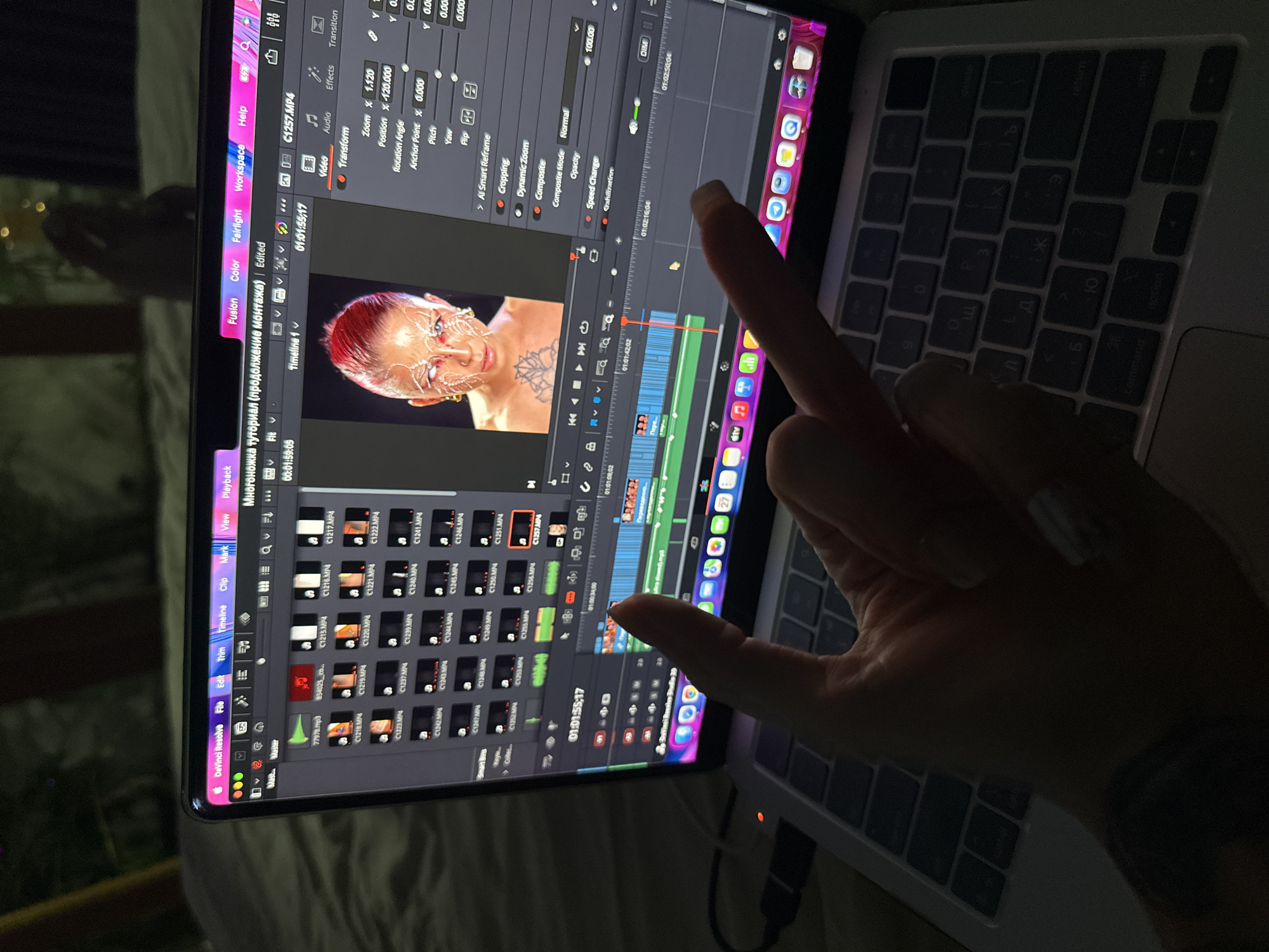 A person's hand points to a laptop screen displaying Davinci Resolve, a video editing software. The software's interface is visible, including the timeline, media pool, and viewer. The project appears to involve a video featuring a person with face paint. The laptop is on a keyboard and is in a dimly lit room. The location is not discernible in the image.