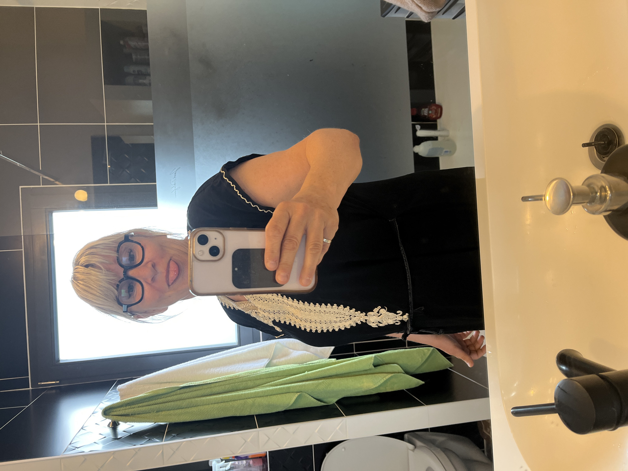 A woman is taking a selfie in a bathroom. She has short blonde hair and is wearing black glasses. She's wearing a black top with a white embroidered design on the front. She is holding a phone in her right hand. The bathroom has black and white tiles, a white sink, and a window. There are towels on the counter. The location is not evident from the provided information.
