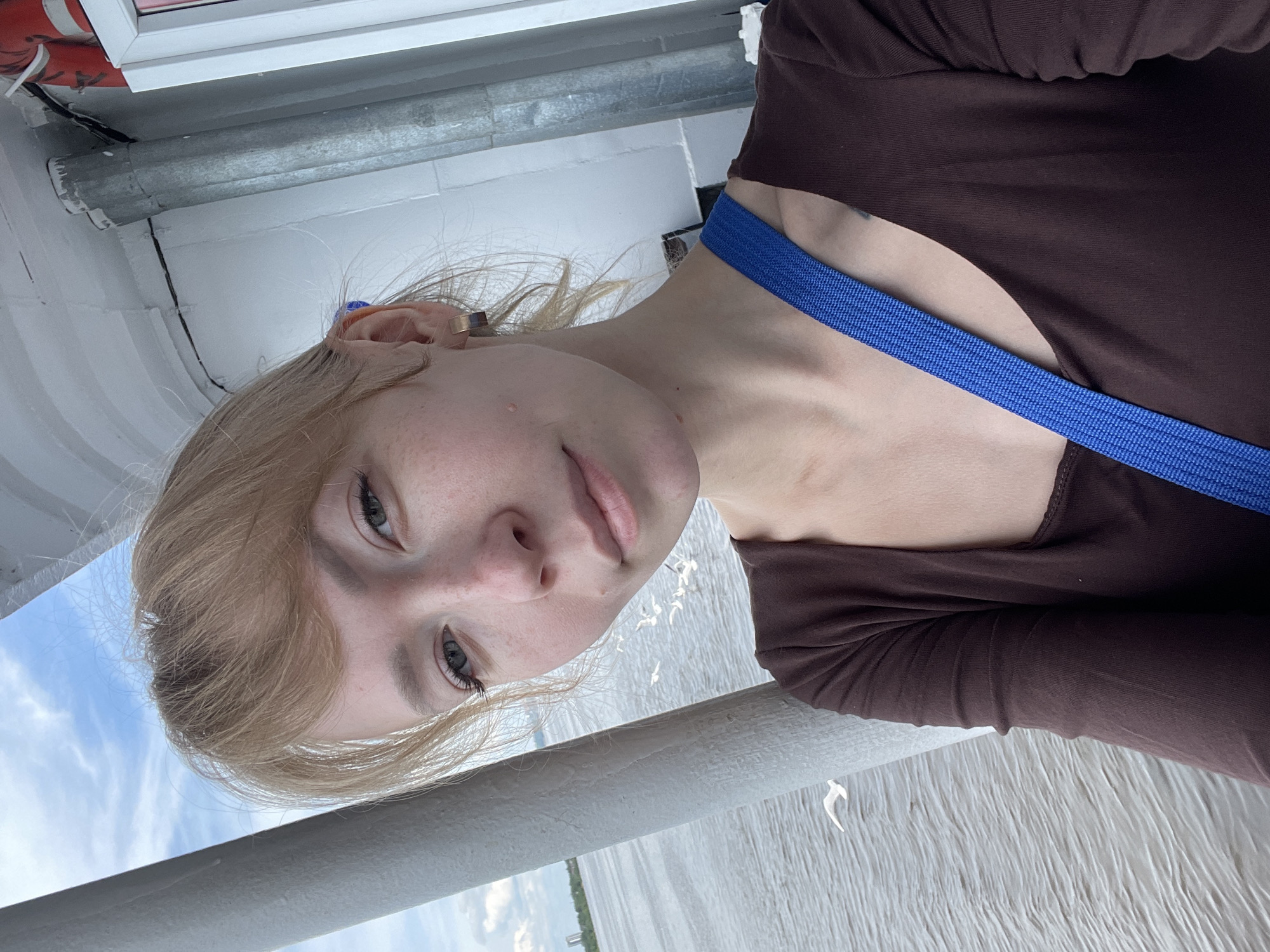 The image is a close-up portrait of a young woman, taken on what appears to be a boat or ferry. She has fair skin, light blonde, chin-length hair with bangs, and light-colored eyes, looking directly at the camera with a neutral, calm expression. She is wearing a dark brown long-sleeved top, with a bright blue strap, possibly from a bag or camera, diagonally crossing her chest. A small, golden ear cuff adorns her right ear.
In the foreground, a light grey railing or bar extends horizontally across the bottom of the frame. Beyond her and the railing, a wide body of water, likely a river, is visible, showing ripples and reflections under natural light. The sky, a clear blue with scattered white clouds, occupies the upper left background. Elements of a white boat structure and additional metal pipes are visible around her.
The scene takes place during the daytime, under bright natural light, suggesting pleasant weather. The setting on a body of water is consistent with Perm, Russia, which is located on the Kama River. No visible text is present in the image.
