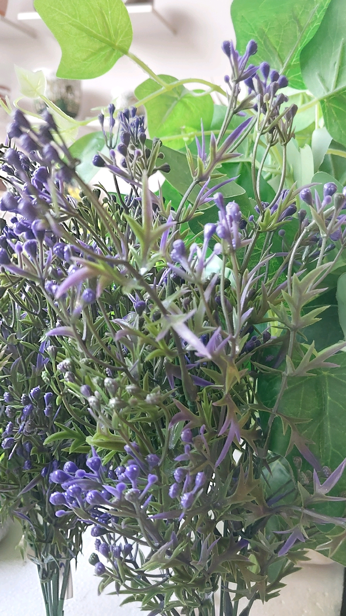 This is a close-up, eye-level, indoor photograph of artificial purple flowers and green leaves. The flowers are small and clustered on thin stems, with some buds visible. The green leaves are broad and ivy-like. The background is blurred, with hints of light and possibly other objects. There are no people visible in the image. The location in Pune, India is not discernible from the image itself.