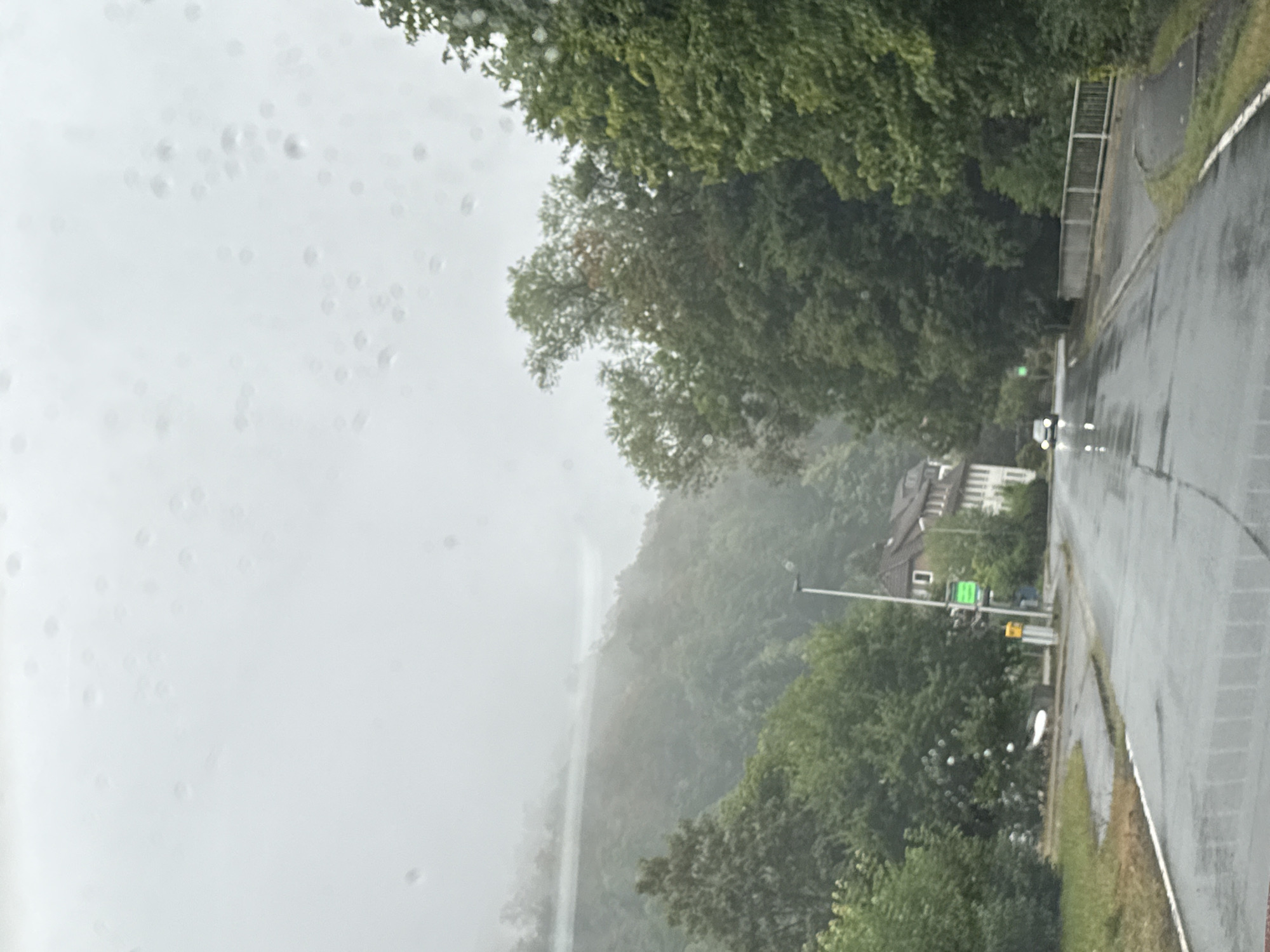 The image shows a rainy day in Walkenried, Germany. A wet road leads into the distance, with trees lining both sides. In the background, there are buildings and a foggy mountain range. A white car is driving on the road. The overall atmosphere is damp and overcast.