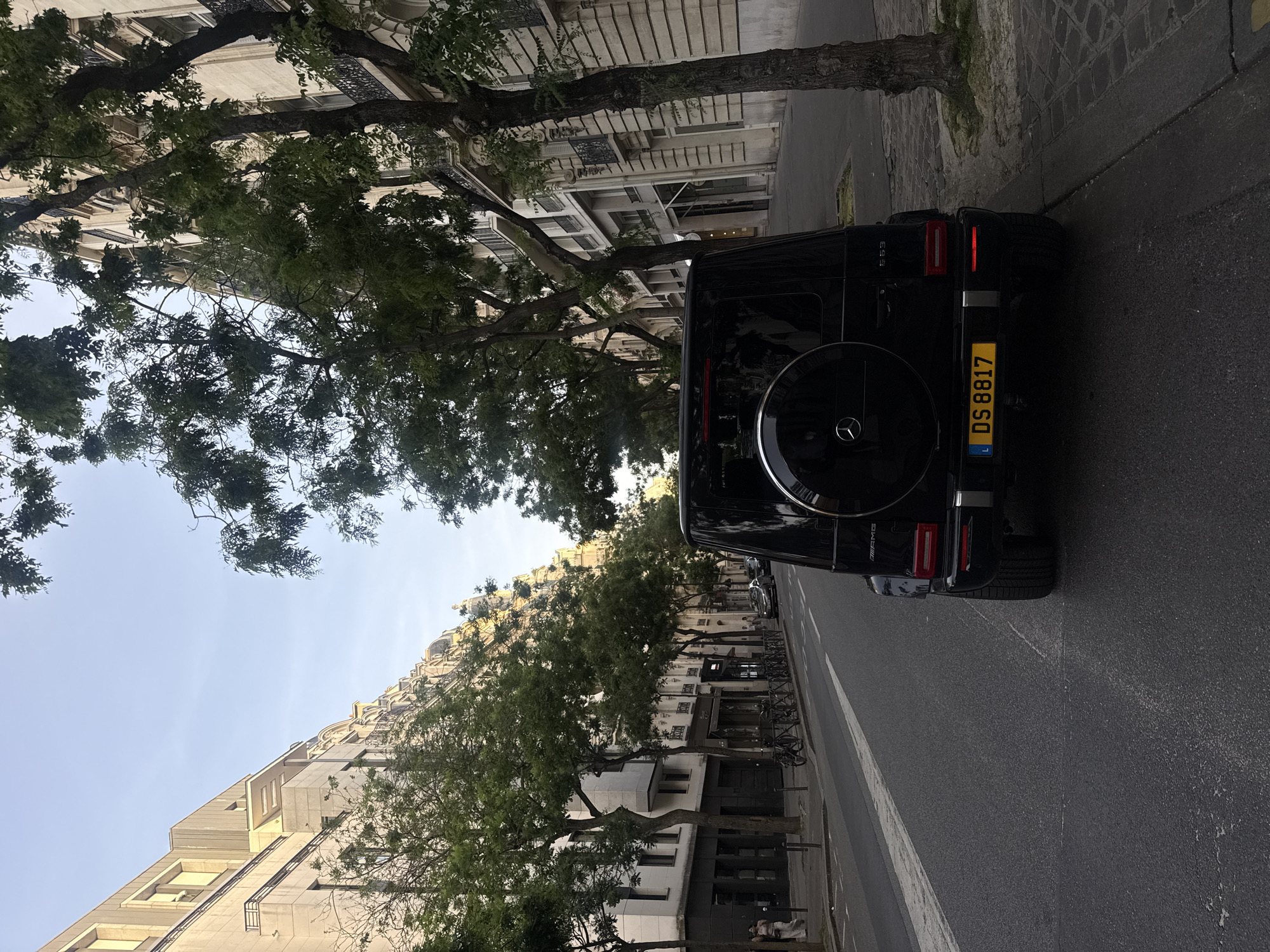 The image depicts a black Mercedes-Benz G-Class AMG parked on a Parisian street. The vehicle, facing away from the viewer, displays a Luxembourg license plate reading "DS 8817". It is parked along the curb, with a paved sidewalk to the right and a single-lane road to the left. The backdrop showcases classic Parisian architecture, featuring beige stone buildings with ornate facades, window details, and wrought-iron balconies. Tall green trees line the street, partially obscuring the buildings. Bicycles are parked along the sidewalk further down the street. The sky is visible in patches through the trees, suggesting either early morning or late afternoon lighting conditions. A person is sitting in front of one of the buildings in the distance. The general ambiance suggests a well-maintained and upscale residential or commercial area in Paris.