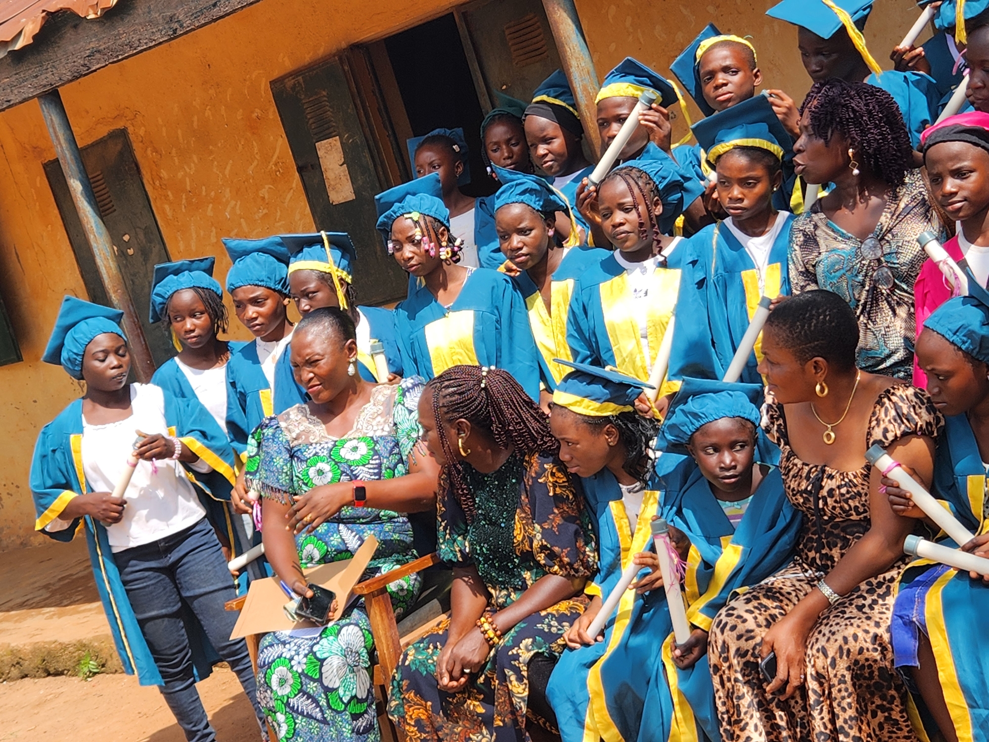 A group of young people wearing graduation gowns and caps are gathered outdoors in front of a building. The building appears to be a school, with a yellow-painted exterior and wooden doors. The students are mostly wearing blue and yellow gowns with yellow caps. Some are holding rolled-up certificates. The scene suggests a graduation ceremony or a related event. The location is identified as Sabon Tasha, Nigeria.