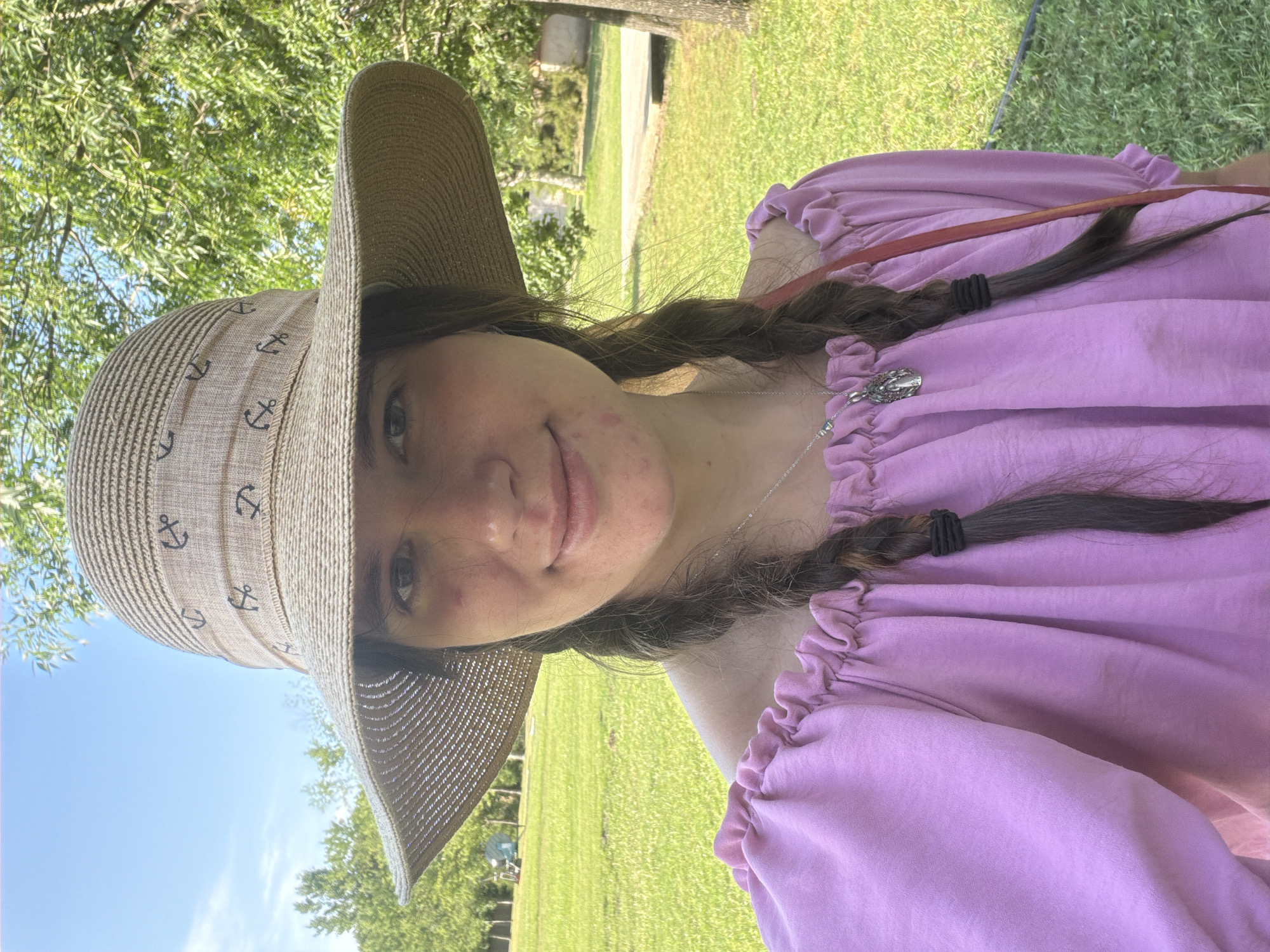 A young woman is taking a selfie outdoors. She is wearing a wide-brimmed, straw hat with an anchor pattern and a ruffled, off-the-shoulder pink top. Her dark hair is in two braids. She is smiling slightly and has some blemishes on her cheek. The background shows a grassy area with trees and a blue sky. The scene appears to be a park or outdoor recreational area in Kiskunmajsa, Hungary.