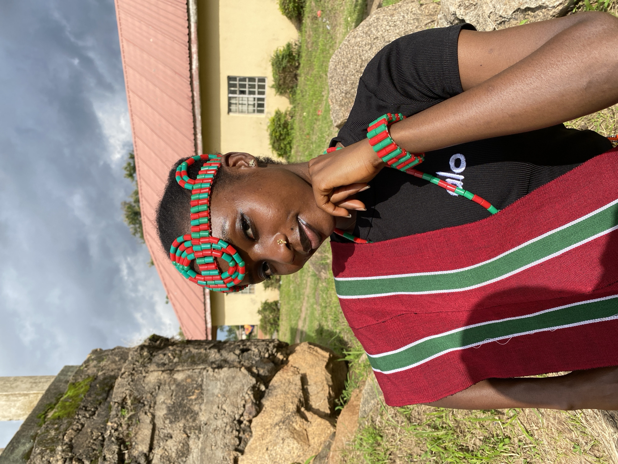 In Wang, Nigeria, a young Black woman, wearing a black t-shirt with the white word "alo" on it, poses for a photo outdoors. She has dark skin, dark hair, and is adorned with a red and green beaded headband and a matching bracelet. A red sash with green and white stripes is draped over her left shoulder. Her right hand rests on her chin, and she is wearing a small gold stud nose ring. The background includes a textured stone structure on the left, grassy terrain, and a yellow building with a red roof and a window on the right. The sky is cloudy.