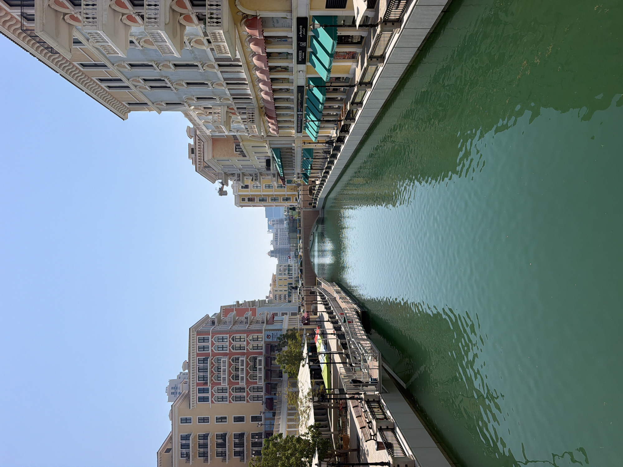 The image captures a sunny day in Venice-inspired architecture along a canal in Doha, Qatar. The scene is dominated by colorful, multi-story buildings with balconies, arched windows, and decorative cornices lining both sides of a tranquil, turquoise canal. On the left bank, there are shops with green awnings and outdoor seating areas with umbrellas and benches. A promenade runs alongside the canal, featuring lampposts and railings. On the right bank, more buildings with similar architectural styles are visible, also featuring shops with green awnings. A bridge can be seen in the distance spanning the canal, with more buildings and a cityscape visible beyond. The sky is clear blue, and the water reflects the buildings and the sky. There are no people prominently visible in the image. The overall impression is one of a well-maintained and picturesque urban environment.