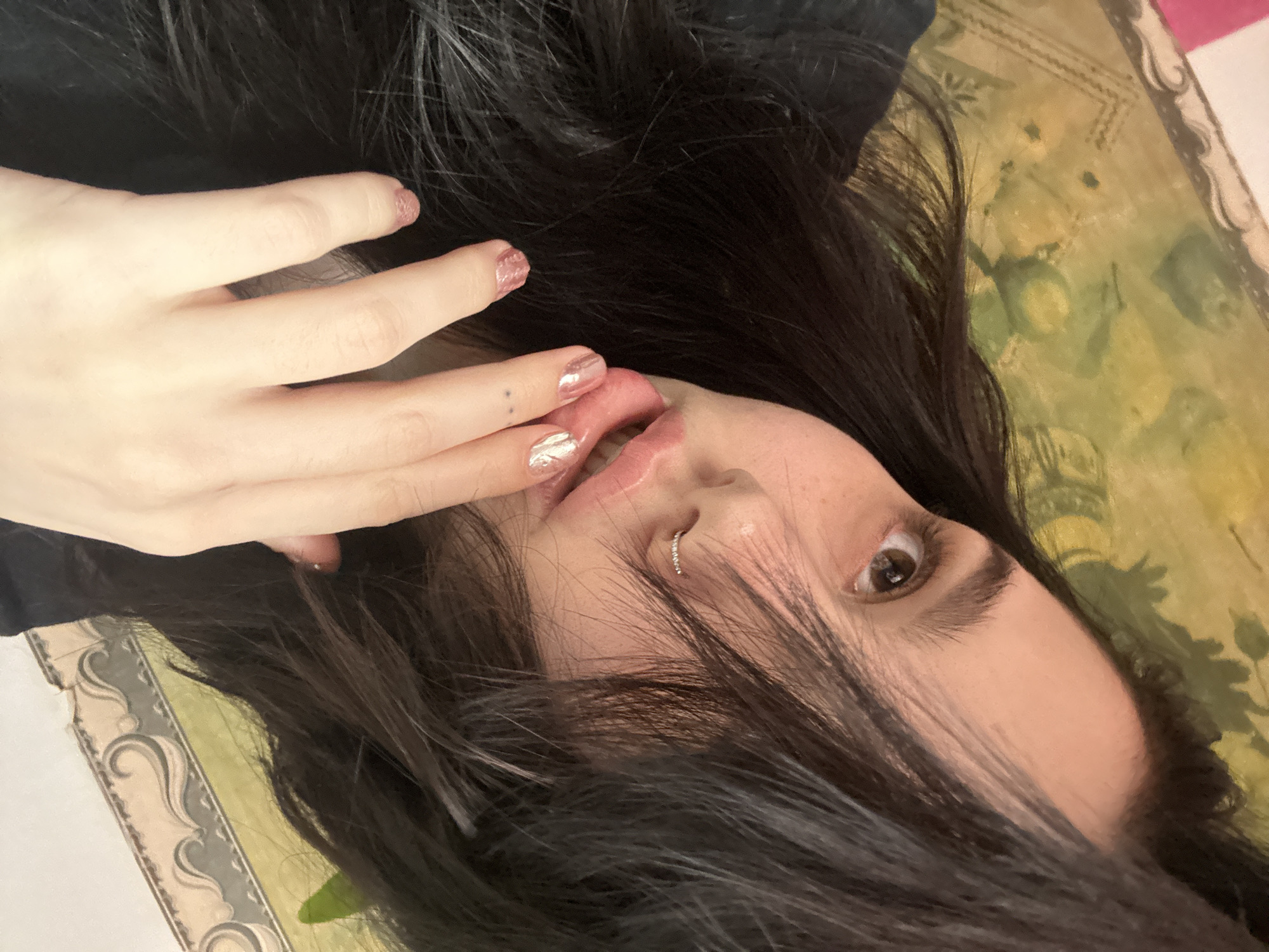 The scene is observed indoors in Aktanysh, Russia. A single individual, identified as female, occupies the majority of the frame, viewed from approximately the chest upwards at an oblique angle. Her long, dark hair is parted and falls forward, partially obscuring her forehead and right eye. Her skin tone is light, with visible freckles on her nose and left cheek. Her right eye, brown in color, is directed towards the viewer. A small, silver-colored hoop piercing is visible on her right nostril. Her lips are slightly parted, revealing no distinct internal features. Her right hand is positioned near her mouth; the index finger, adorned with glittery, light pink nail polish, is lightly touching the lower lip. The other visible fingers of the right hand also feature this same nail polish. A small, dark tattoo, appearing as multiple small dots, is located on the back of her right middle finger, near the knuckle. She wears a dark garment, possibly a t-shirt. The background consists of a patterned surface, likely a wall covering or fabric. To the left, a faded yellowish-green pattern with abstract or floral elements is present. To the right, a lighter section with a decorative border featuring grey scrollwork is visible. A small section of a vibrant pink object is visible at the extreme bottom-left edge of the frame.