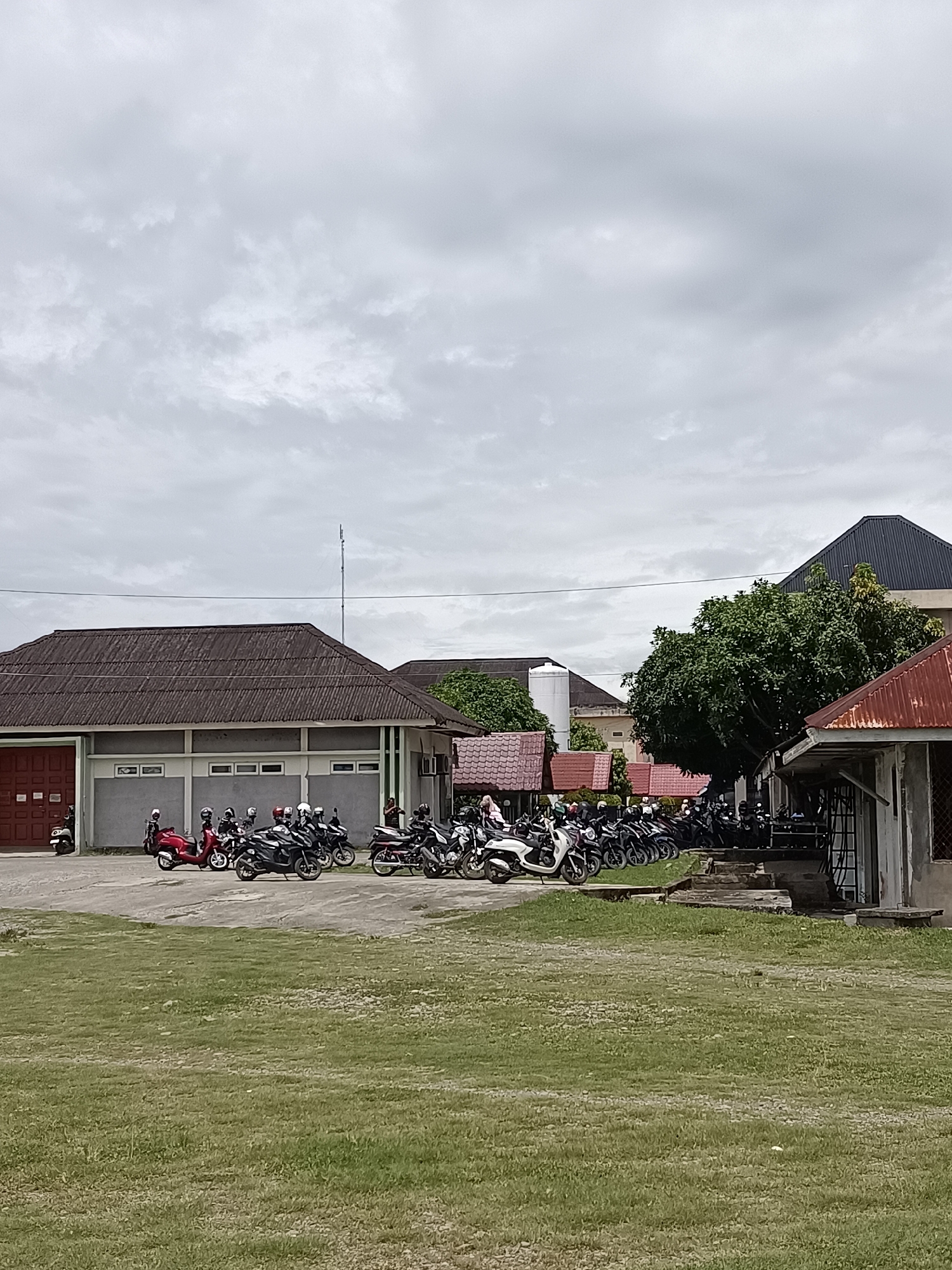 The image captures a cloudy day scene in Mesjid Reuntoh, Indonesia, featuring a parking lot filled with numerous motorcycles in front of several buildings. The foreground is dominated by a large, flat expanse of green grass, suggesting an outdoor public space or a park.
In the midground, a paved area serves as a parking lot. It is densely populated with a variety of motorcycles, most of which appear to be scooters or smaller commuter bikes. Many of the motorcycles have helmets on their handlebars or seats, indicating recent use. Several people, dressed in casual clothing, are visible among the motorcycles, some standing and others seemingly in the process of parking or preparing to leave.
Behind the parking lot, several structures are present. The most prominent building on the left has a long, dark tiled roof and a reddish-brown double door. It appears to be a functional building, possibly an office or a community center, with a row of rectangular windows along its facade. To the right of this building, a line of smaller structures with reddish-brown, corrugated metal roofs are visible, suggesting a more informal arrangement of buildings or covered areas. Further to the right, a larger building with a dark, angular roof is partially obscured by a large, leafy green tree. Another smaller, rustic structure with a rusted metal roof stands in the far right of the midground, adjacent to the larger building.
The sky is overcast with diffused grey clouds, creating a soft, even light across the scene. The overall impression is one of a busy but orderly outdoor environment on a day with moderate weather conditions.