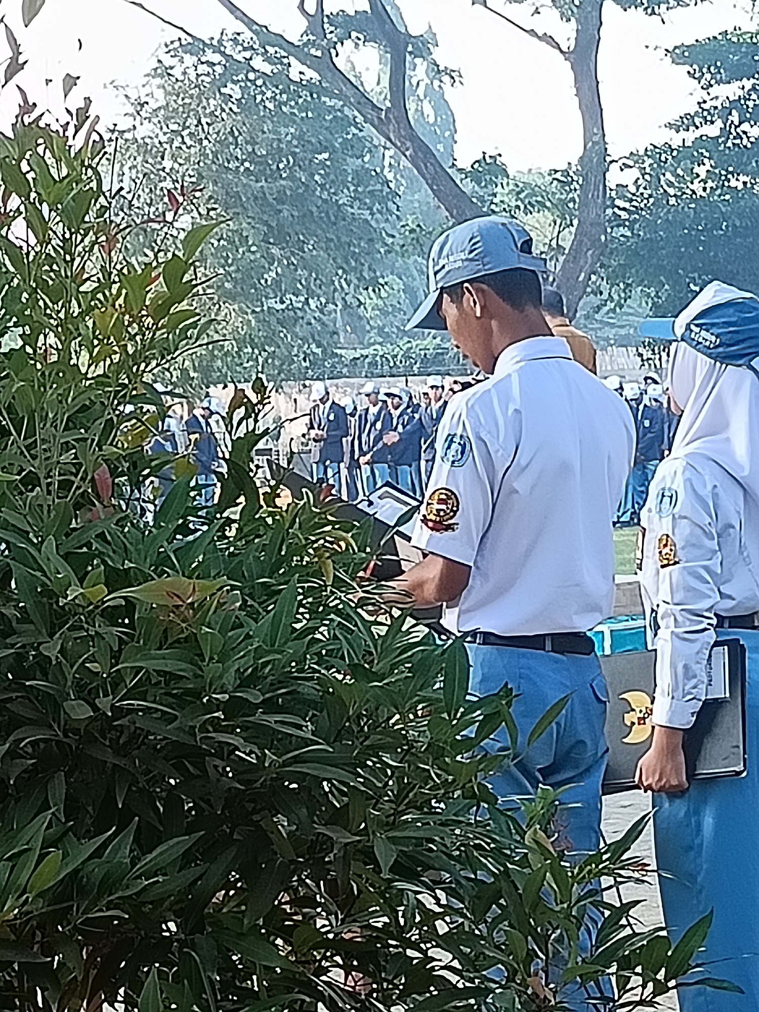 In Kediri City, Indonesia, two students in white shirts and blue pants, likely on a school field trip or event, stand near a bush. The male student on the left wears a blue cap and is looking down at a book or document. The female student on the right wears a white hijab and is holding a folder. In the background, a group of students in similar uniforms are gathered, with trees and a fence suggesting an outdoor setting.