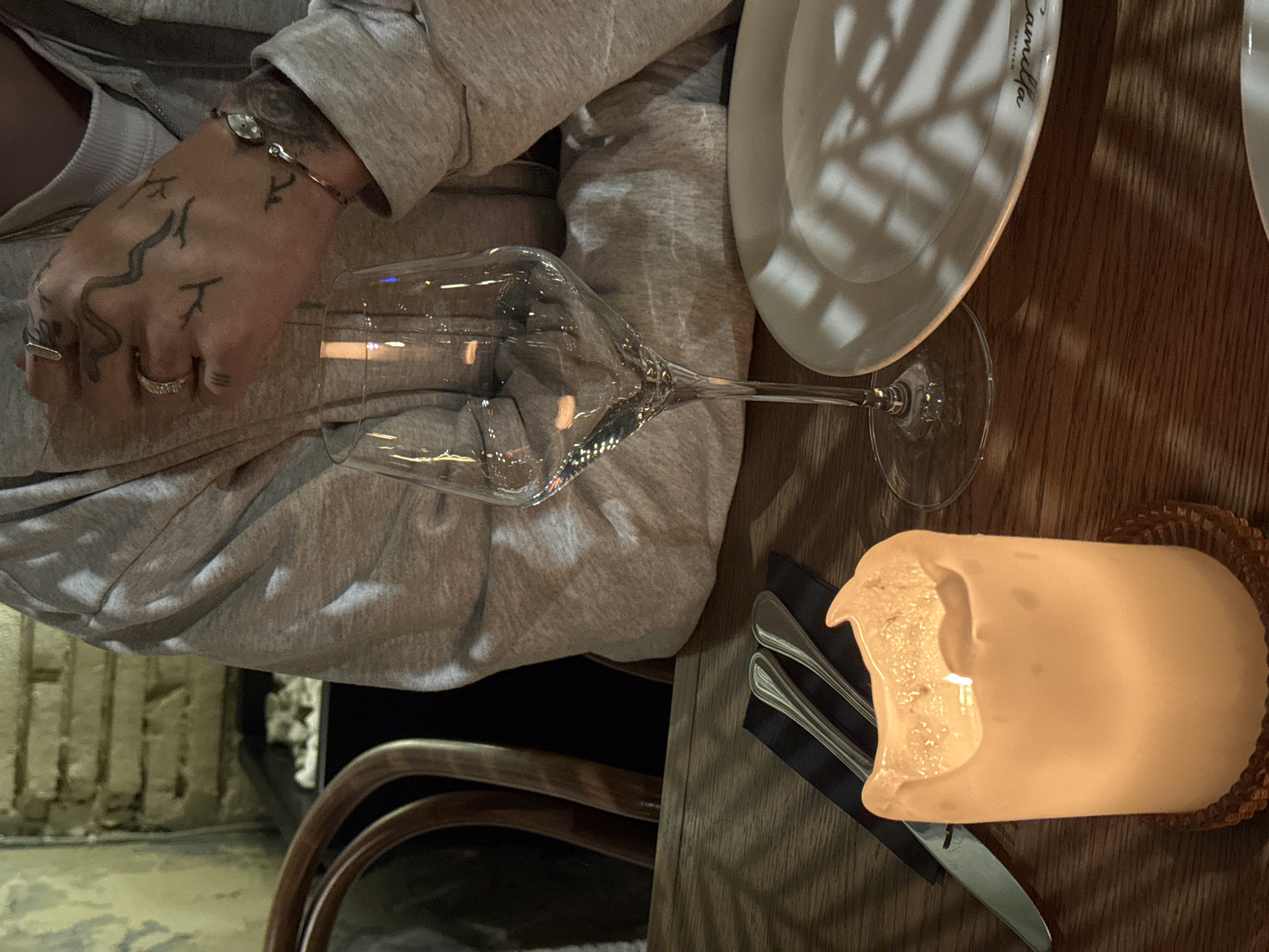 A dimly lit, eye-level shot reveals a table setting in Yerevan, Armenia, with a hand resting near a wine glass. The hand is adorned with rings and tattoos. A white plate, a lit candle in a wicker holder, and cutlery are arranged on the wooden table. A partial view of a chair and textured wall can be seen in the background.