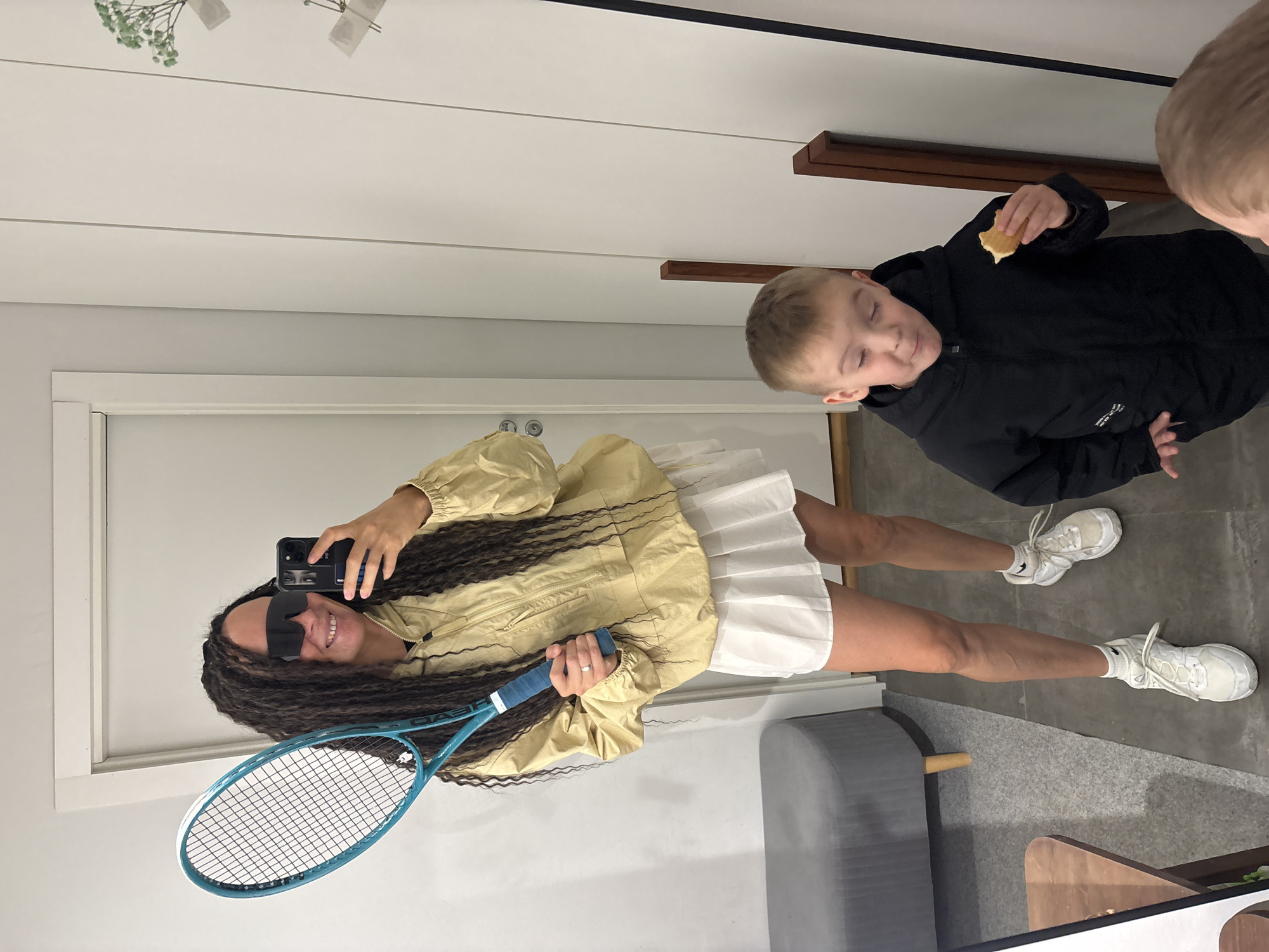 A woman with dark, long hair holds a tennis racket and a mobile phone up, facing a mirror. She is wearing sunglasses, a yellow jacket, and a white tennis skirt with white sneakers. A young boy in a black jacket is on the right, eating something. Another child's head is partially visible above him. The background shows a white wall, a gray stool, and a wooden shelf. There is no indication of the location being Chaikovsky, Russia.