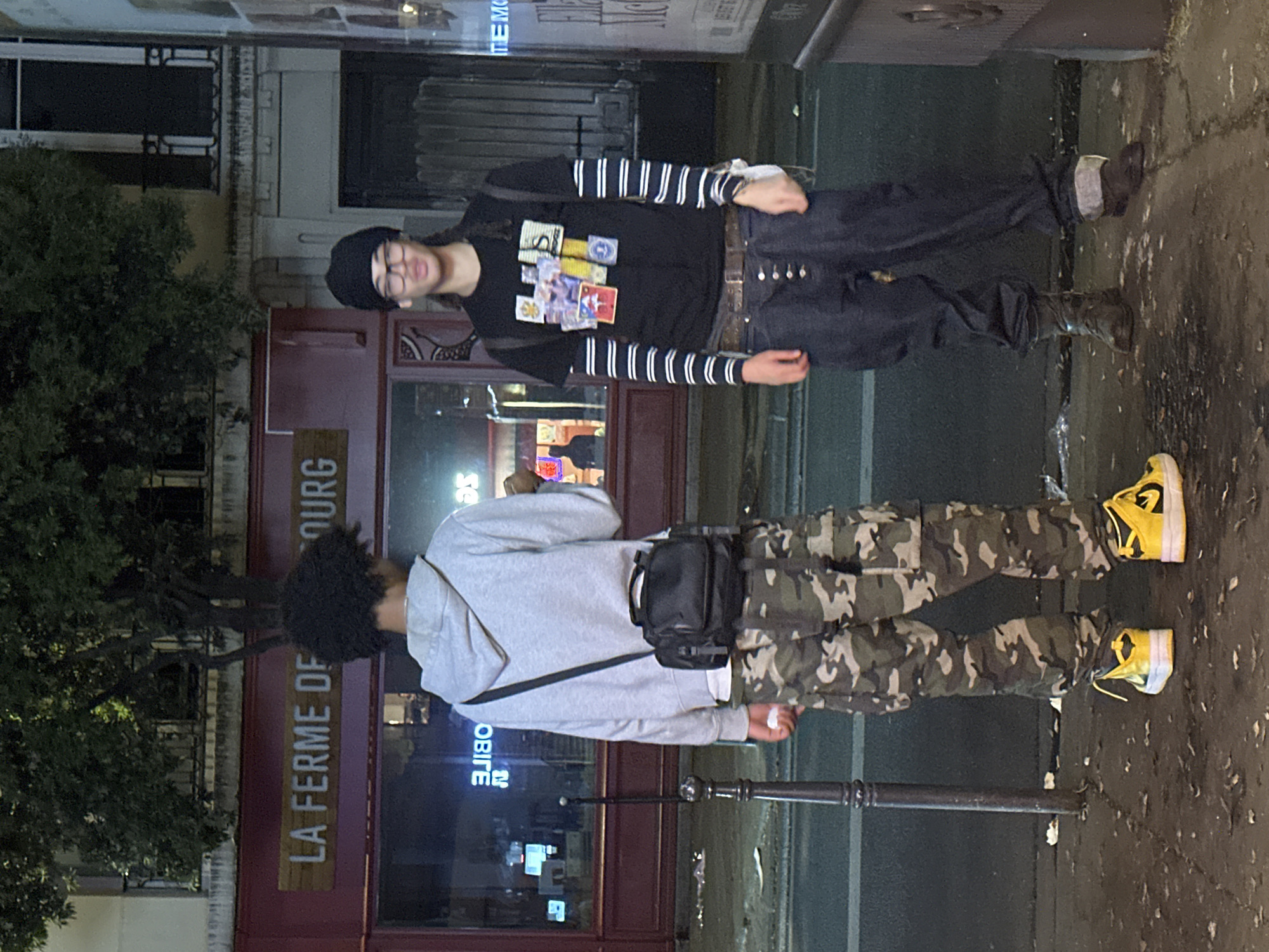 This photograph captures two individuals standing on a wet urban street at night in Paris, France. The ground is dark and reflective, suggesting recent rain. Artificial light sources illuminate the scene, including streetlights and reflections from building interiors.
The person on the left is a young individual facing the camera, wearing a black beanie and glasses, with a slight smile. They are dressed in a black t-shirt featuring colorful patches or graphics, layered over a long-sleeved shirt with horizontal black and white stripes. They wear dark pants and dark boots.
The second individual, positioned to the right, has their back to the camera. They are wearing a grey hooded sweatshirt, camouflage pants, and distinct yellow and white sneakers. A black shoulder bag is slung across their chest. Dark, curly hair is visible.
In the background, a building with a reddish-brown facade is prominent, displaying the French text "LA FERME DU BOURG" on a wooden sign. Other faint, reversed text such as "OBITE" (likely part of "MOBILE") and "MEMO" can be seen on various parts of the buildings. A dark street pole stands between the two people, and a tree is visible on the far left. The overall setting is a casual street scene at night.