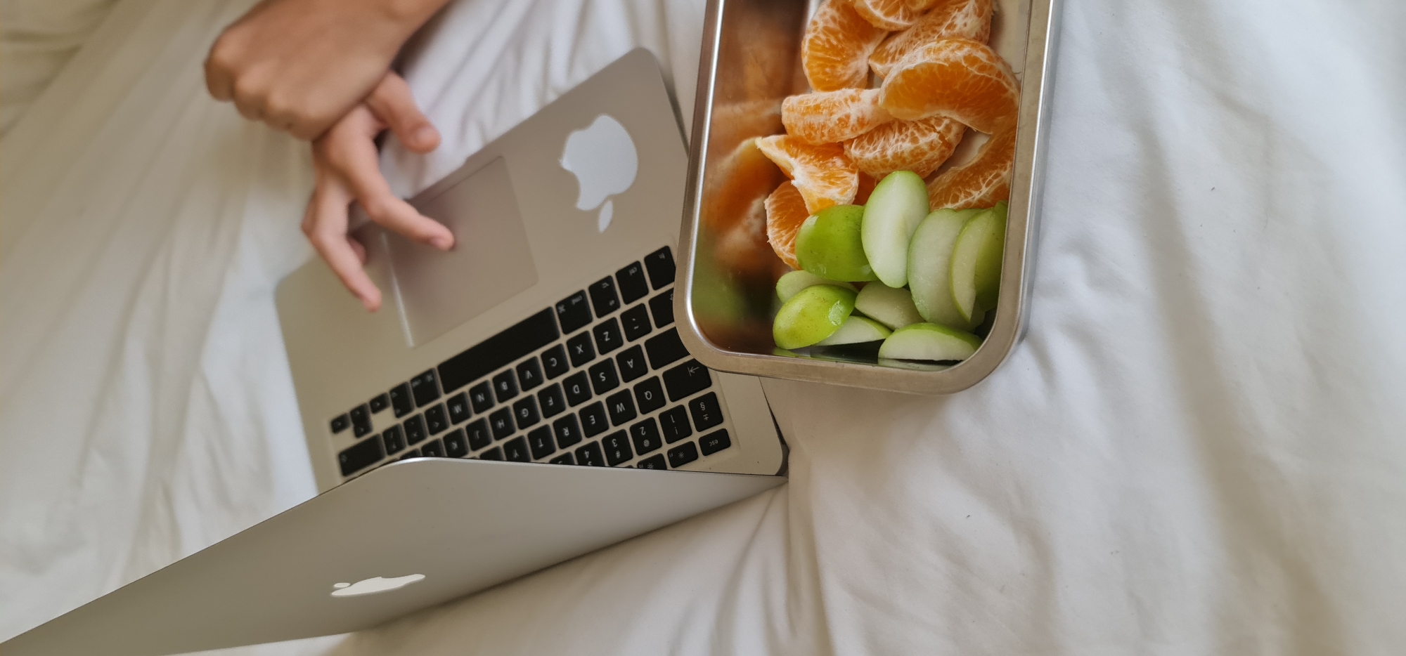 The image shows a person using a silver laptop on a white bed. Beside the laptop is a silver rectangular container filled with slices of oranges and green apples. The person's hand is resting on the trackpad of the laptop. The environment appears to be indoors on a bed with white linens.