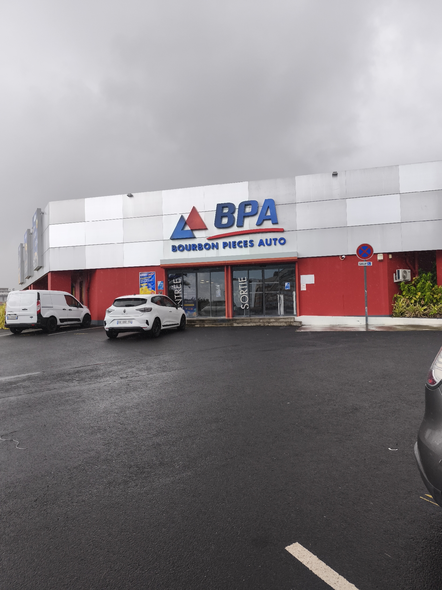 The image shows the exterior of a BPA (Bourbon Pièces Auto) store located in Saint-Denis, France. The building features a red lower facade with white paneling above, displaying the blue BPA logo and the text "BOURBON PIECES AUTO". Two white vehicles are parked in the paved lot in front of the store: a white van on the left and a white hatchback on the right. The entrance and exit doors are visible, with signage indicating "ENTRÉE" (entrance) and "SORTIE" (exit). A red and white circular sign with a blue symbol is mounted on a pole to the right of the entrance. The ground is wet, suggesting recent rain, and the sky is overcast. Part of a grey car is visible in the lower right corner of the image.