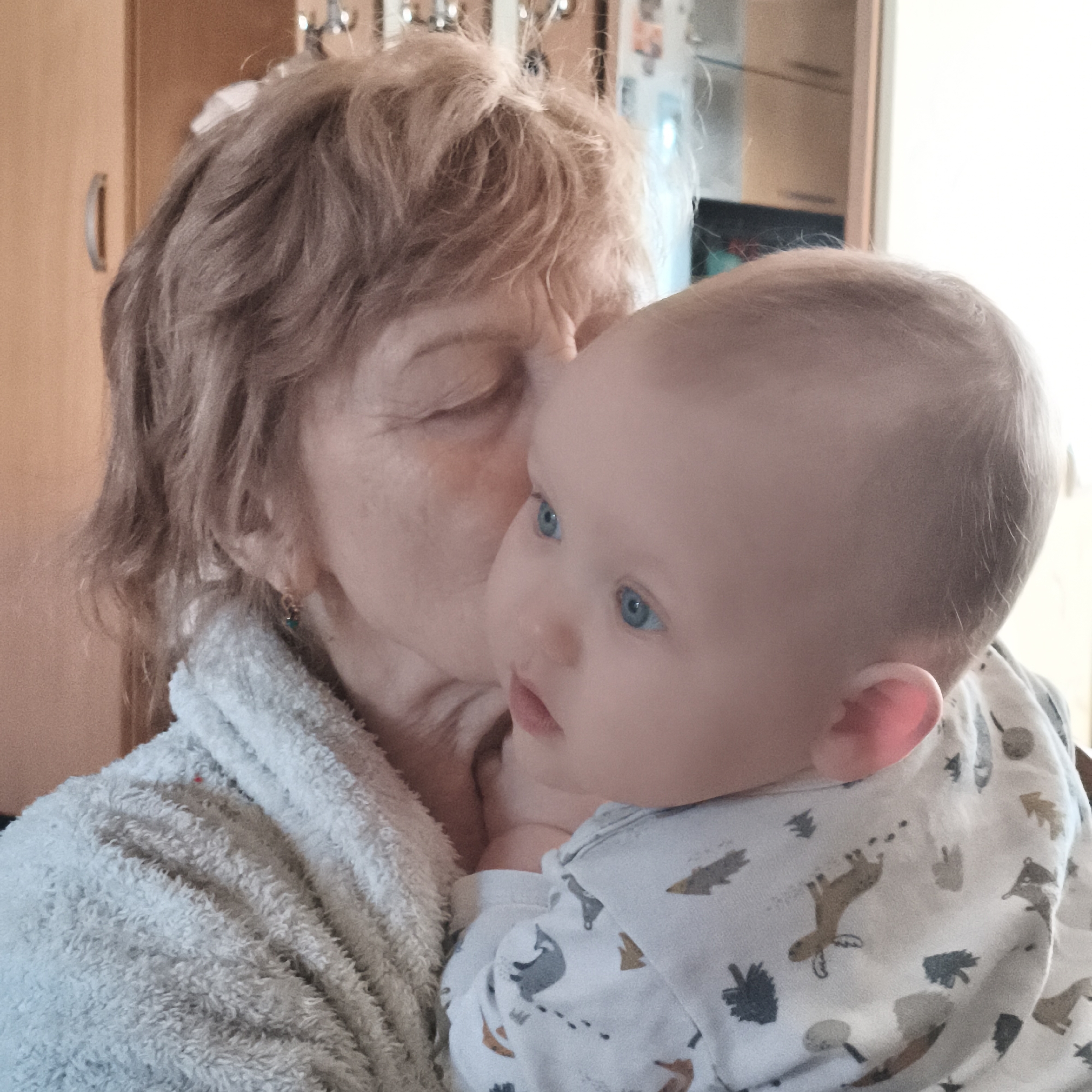 This intimate indoor photograph, taken in Chaikovsky, Russia, captures a tender moment between an adult woman and a baby. The woman, who appears to be an older adult with light brown, wavy hair, is shown from the chest up, wearing a light-colored, fluffy robe. A small, golden earring with a blue-green bead is visible in her right ear. Her eyes are closed as she gently kisses the baby on its temple or cheek, conveying deep affection and tenderness.
The baby, with fair skin, very fine light hair, and striking blue eyes, is held in the woman's arms. It is dressed in a white long-sleeved onesie or sleepsuit patterned with cartoonish forest animals like deer, trees, and mushrooms. The baby is looking towards the right side of the frame with a neutral expression.
The background, softly blurred, indicates an indoor home setting with warm-toned wooden cabinets or furniture visible. The natural, soft lighting suggests it is daytime. The image beautifully encapsulates a loving interaction between generations. There is no visible text in the media file.