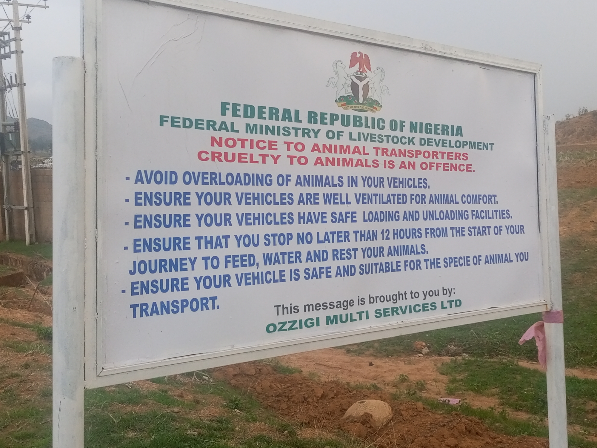 The image depicts a sign in Kuru Dungus, Nigeria, bearing the official coat of arms of the Federal Republic of Nigeria. The sign is a public notice from the Federal Ministry of Livestock Development aimed at animal transporters. It advises against overloading animals, ensures vehicles are ventilated, have safe loading facilities, and that drivers stop every 12 hours to feed, water, and rest the animals. The sign also stresses that vehicles should be safe and suitable for the specific animals being transported. The message is attributed to OZZIGI MULTI SERVICES LTD. The sign itself is white with blue and red text. The surrounding environment appears to be a rural or semi-rural area with bare earth, sparse vegetation, and utility poles in the background.