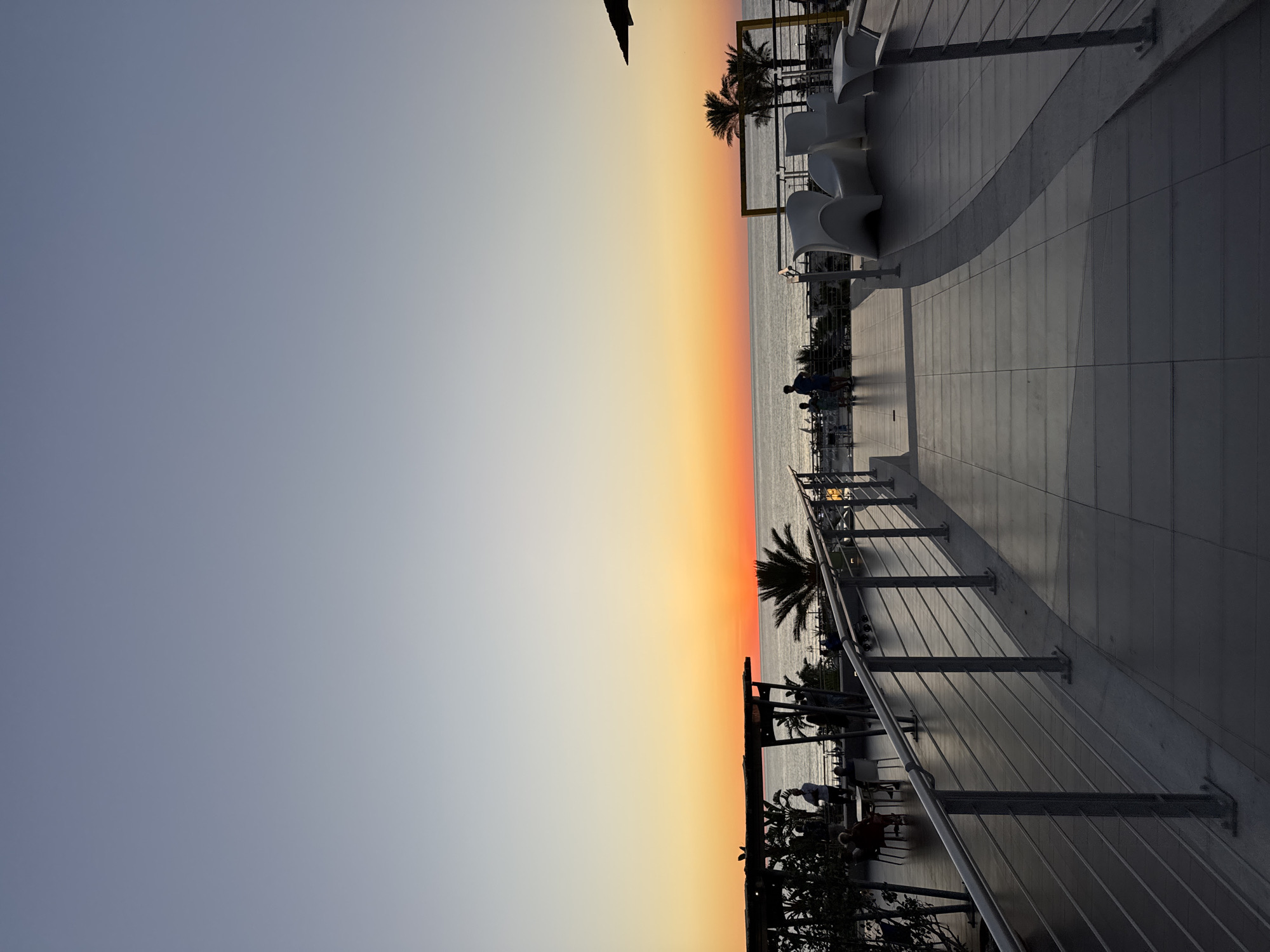 The image shows a sunset over the Mediterranean Sea in Isola delle Femmine, Italy. The sky transitions from a pale blue at the top to vibrant orange and red hues near the horizon. A body of water, likely the sea, is visible. In the foreground, a paved walkway with a metal railing leads towards the sea. On the walkway, several white modern chairs and a table are arranged. To the left, there's a covered seating area with tables and chairs, where several people are seated and standing. Palm trees and other foliage are present. Two figures, silhouetted against the sunset, stand on a lower level overlooking the sea. The overall scene depicts a tranquil evening at a coastal venue.
