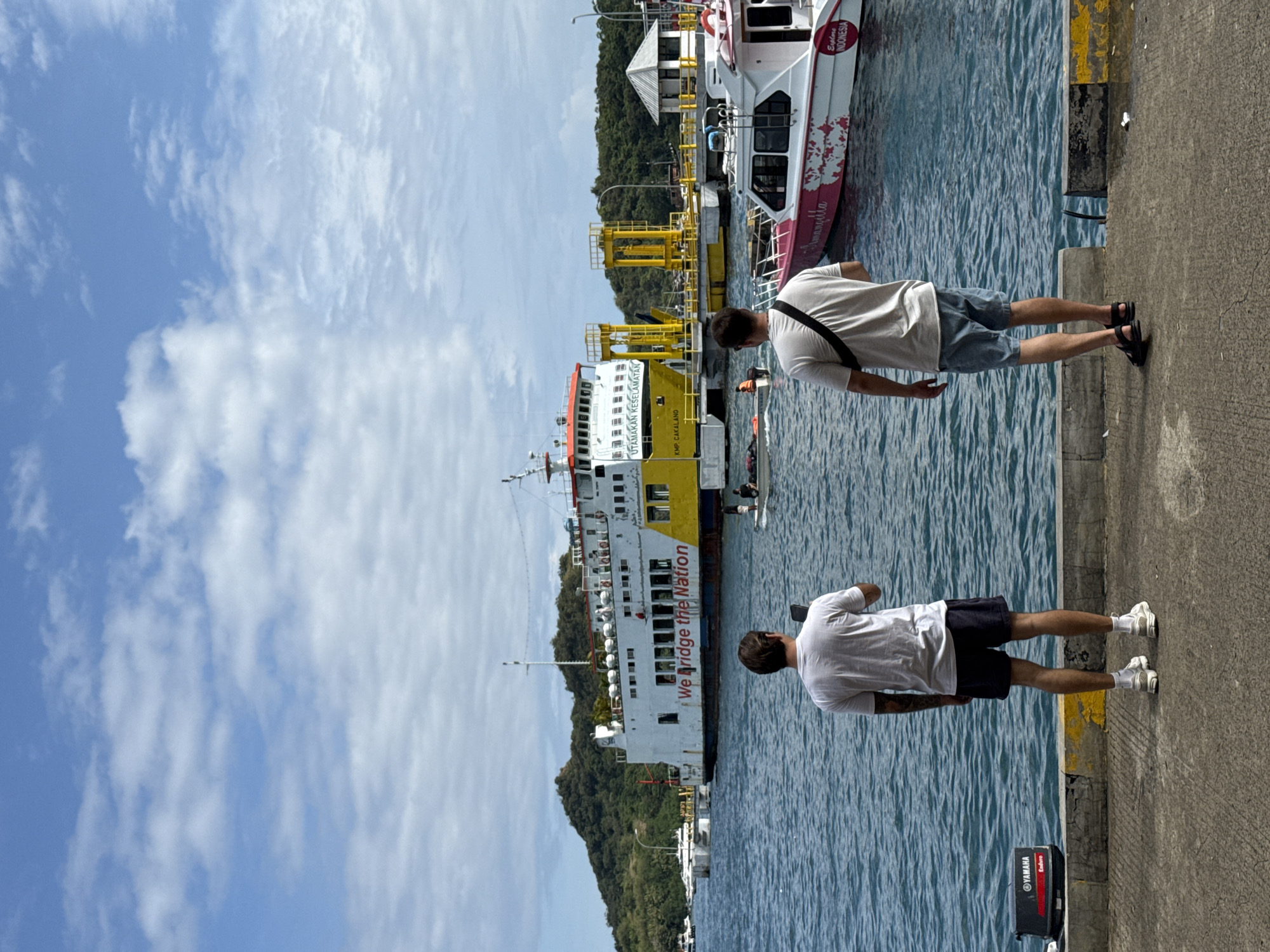 The image captures a scene at the harbor in Labuan Bajo, Indonesia. Two men, each wearing a white t-shirt and shorts, stand facing the water along the concrete edge of the harbor. One man wears blue shorts and black sandals, while the other wears black shorts and white sneakers. A large ferry boat, predominantly white with yellow sections, is docked nearby. Red lettering on the side of the boat reads "We Bridge the Nation." Another boat, pink and white, is docked closer to the foreground. The water in the harbor reflects the sky. A green, hilly landscape forms the backdrop. A clear sky with scattered clouds is visible overhead. Near the concrete edge, a black Yamaha outboard motor box is also visible.