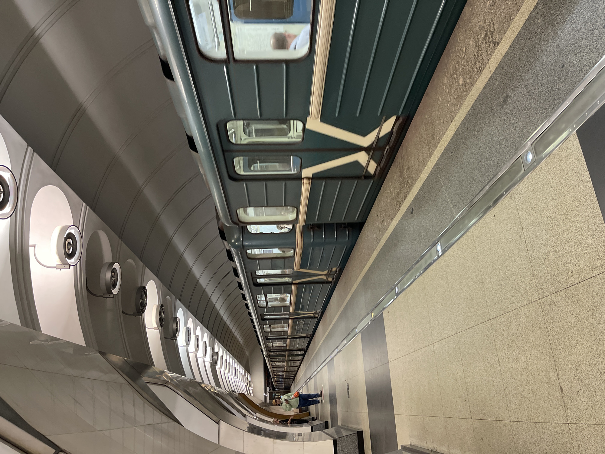 The photograph shows an underground subway station. A train is visible from the side, parked at the platform. The subway car is dark green with white stripes. Windows are visible and reflect the interior of the station. The platform is made of tan-colored, square tiles. A person stands on the platform. The wall of the station is arched and adorned with rows of circular light fixtures. The lights have a black ring on the outside. The station is modern. The location is unknown.
