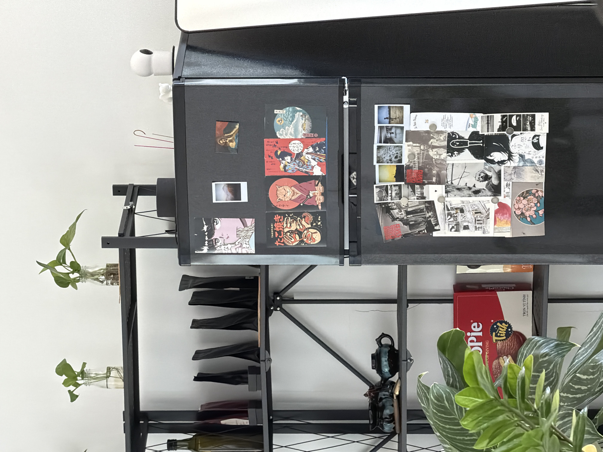 A shelf with various items is centered in the frame. The shelf has black metal supports with a criss-cross pattern near the bottom. On the left side, plants in glass jars are attached to the shelf. A wine rack holds bottles, while other decorative objects, including what looks like a camera, are located on other levels. Boxes of "Chocopie" are on the shelf, as well as the spines of books. The refrigerator above the shelf is covered in magnets, polaroids, and cards. The text on the "Chocopie" box mentions "Đà Nẵng, Vietnam".