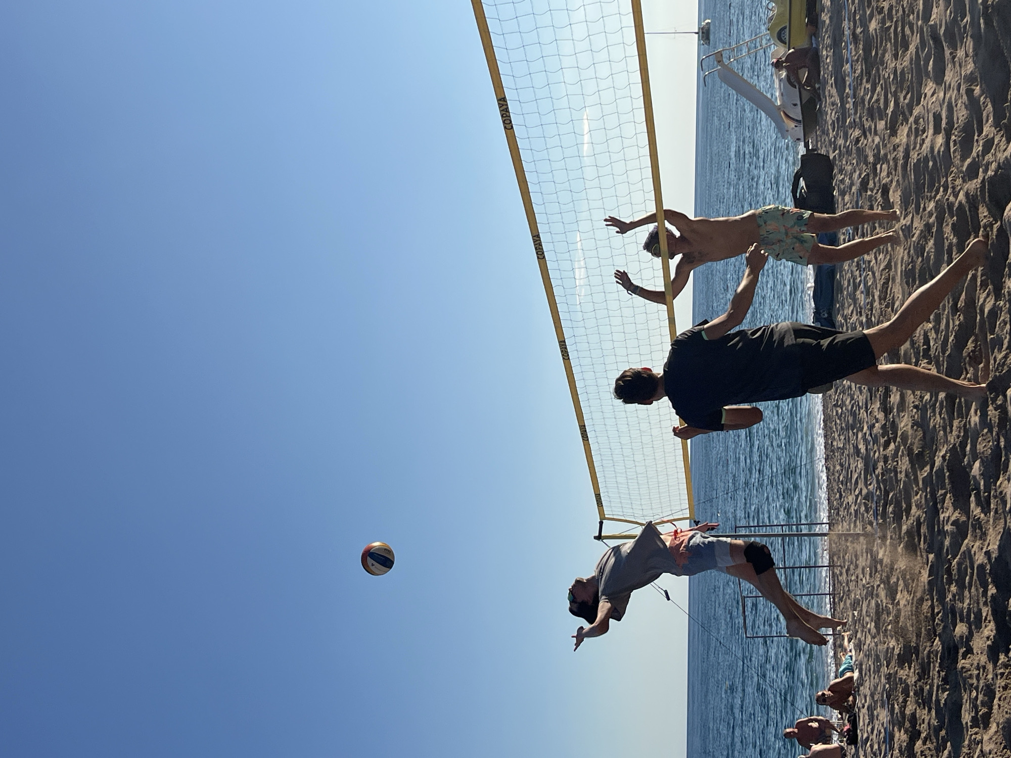 A group of young men are playing beach volleyball on a sandy beach next to the sea. The sky is clear and blue. A volleyball net is set up, and one man is jumping to spike the ball while another prepares to receive it. Two other men are also visible near the net, one in the foreground and one in the background. Several other people are sitting and relaxing on the beach in the background. A few beach chairs, umbrellas, and some equipment are scattered on the sand. The water appears calm, and a few sailboats are visible in the distance. The overall scene depicts a lively recreational activity on a sunny day at the beach.