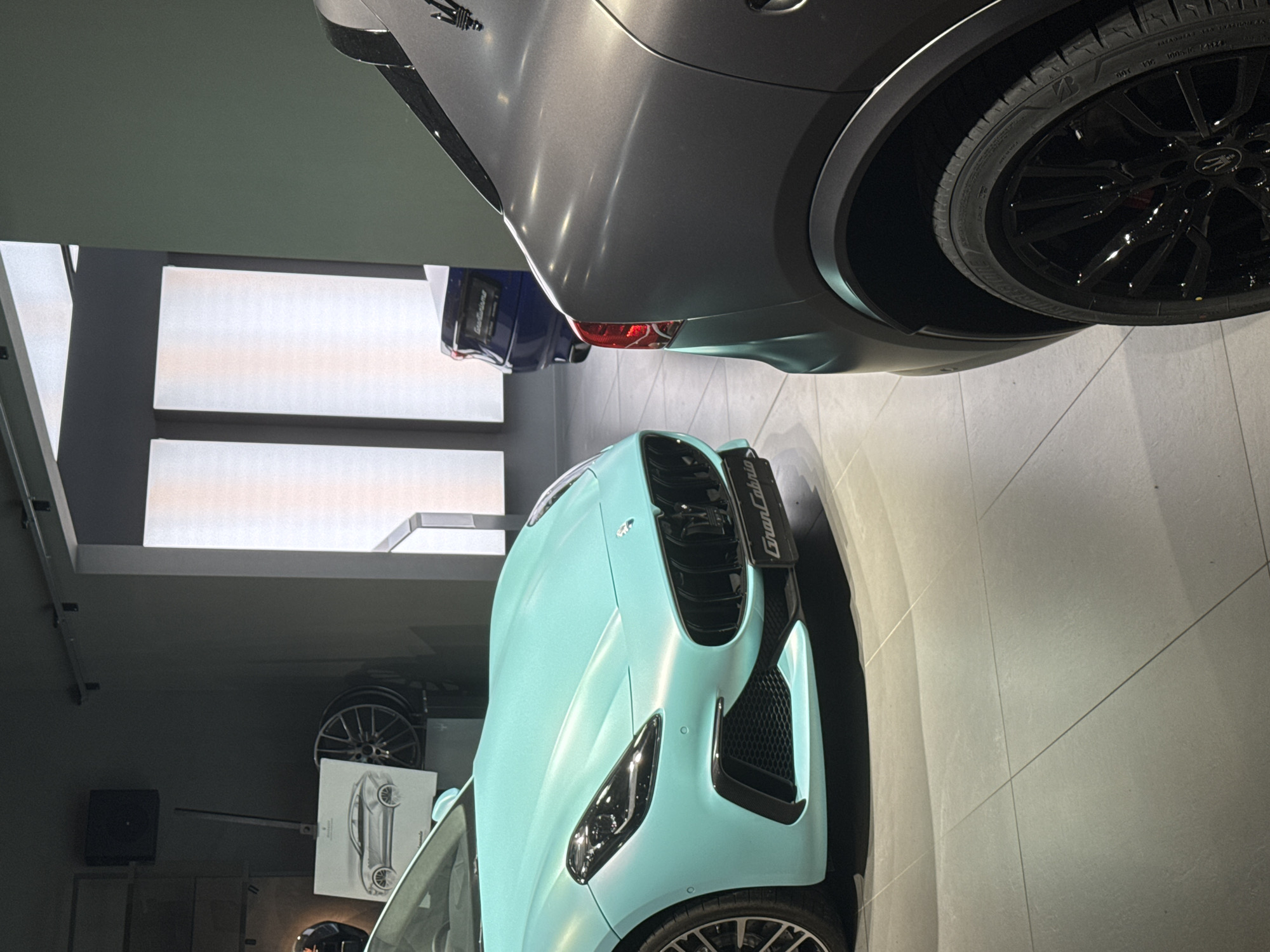 The image shows the interior of what appears to be a car dealership or showroom. Dominating the view are three Maserati vehicles: a light blue GranCabrio convertible in the foreground, a dark grey sedan in the upper right, and a glimpse of a blue car further back. The location seems to be a well-lit, modern space with large windows and tiled floors. In the background, there's a display featuring car wheels and a framed technical drawing of a car. I do not have information about specific locations in Sofia, Bulgaria.