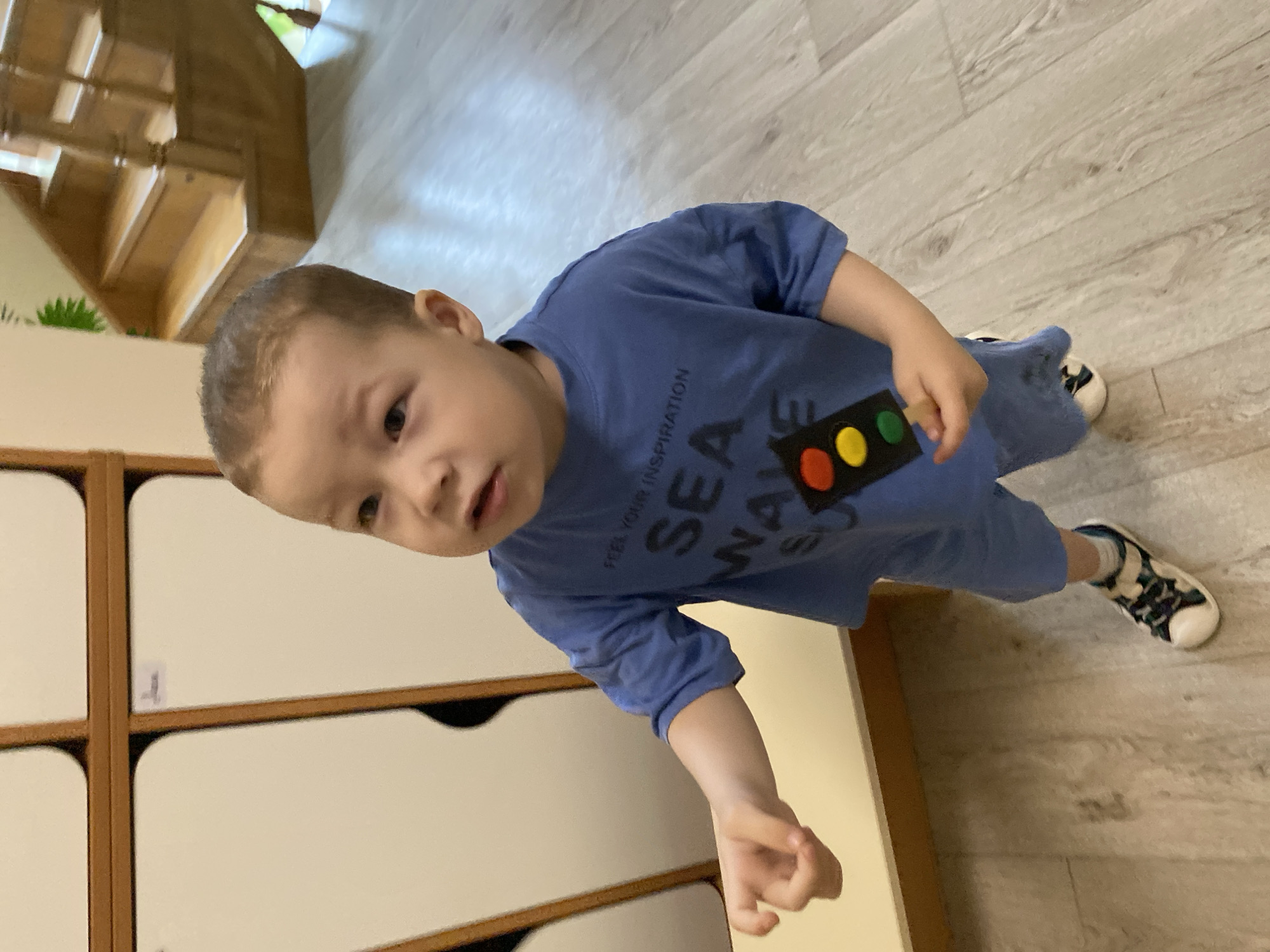 A young boy stands in a room with light-colored wooden floors. He wears a blue t-shirt with a traffic light emblem and blue shorts, and black-and-white sneakers. He is positioned near a set of white cabinets with wood trim. The background includes a wooden staircase. There is no evidence or indication that this is in Sochi, Russia.