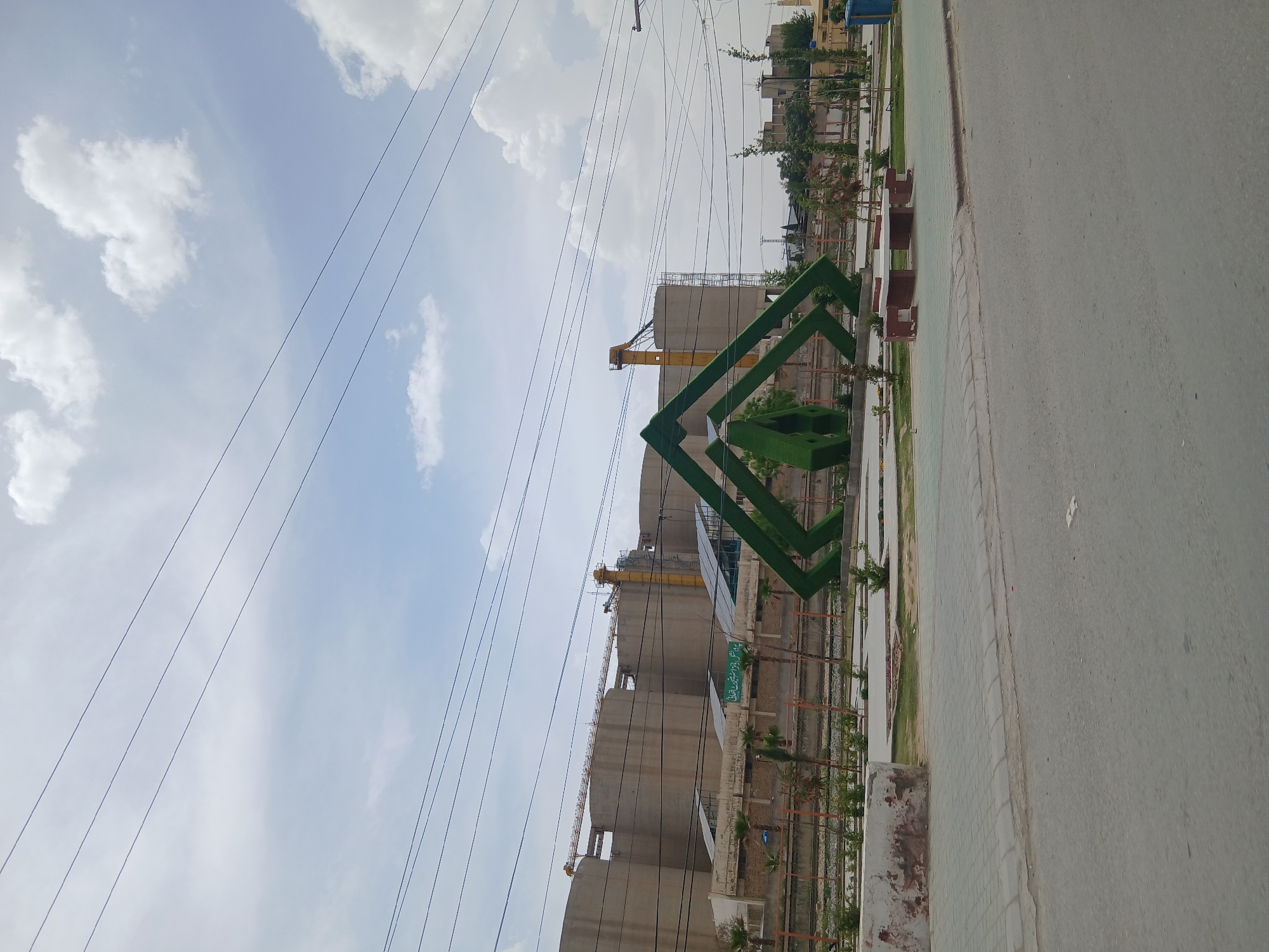 The image captures an outdoor scene in Quetta City Tehsil, Pakistan, under a partly cloudy sky. Numerous overhead power lines stretch diagonally across the upper portion of the frame.
In the foreground on the right, a paved road runs alongside a concrete sidewalk and a landscaped strip. This strip features a decorative green structure composed of interconnected geometric shapes, some of which appear to be covered in artificial grass. Near this structure, concrete benches are visible among small plants and shrubs, suggesting a public amenity area.
The midground is dominated by several large, cylindrical concrete silos or storage tanks, indicative of an industrial facility or a large-scale construction site. A bright yellow crane is positioned between two of these silos, suggesting ongoing heavy construction or industrial activity. A green sign with white text, likely in Urdu, is attached to one of the silos. Portions of the text visible include "صوبائی سیکرٹیریٹ" (Provincial Secretariat) and potentially "کالا پانی" (Kala Pani).
Further in the background, other buildings and structures can be discerned, hinting at a developed urban or suburban area. No people are visible in the frame. The scene is brightly lit by daylight, suggesting it is daytime under clear to partly cloudy weather.