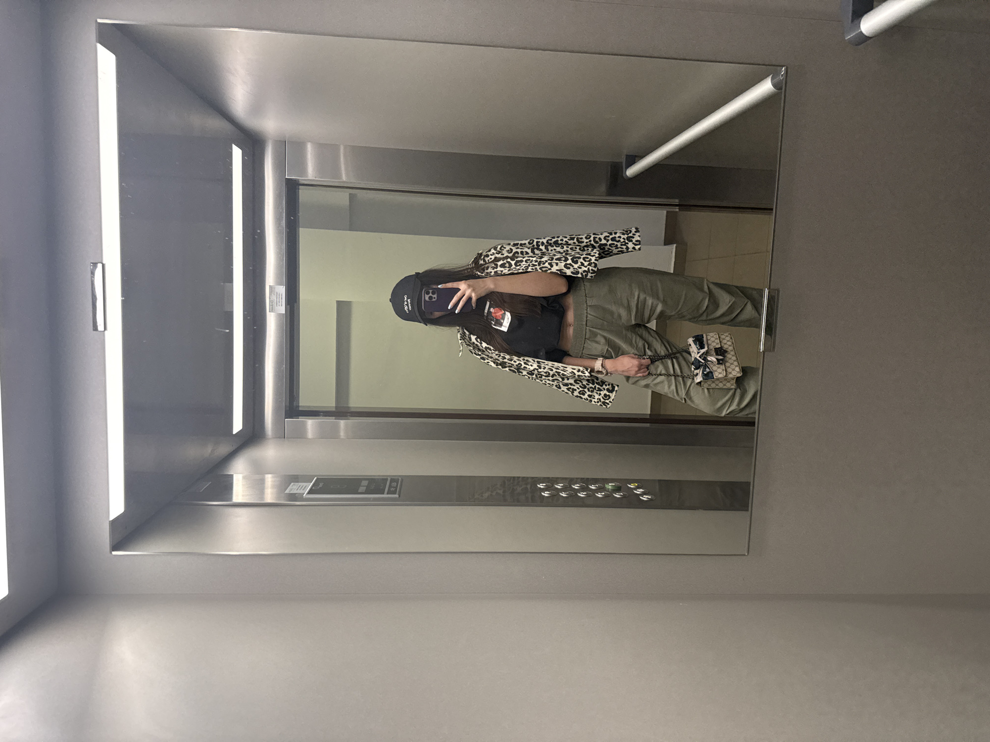 The image shows a person taking a selfie in a mirror inside an elevator. The person is wearing a black t-shirt, olive-green pants, a leopard-print jacket, and a black baseball cap. They are holding a smartphone, presumably taking the photograph. The reflection shows the interior of the elevator, including the button panel and the opposite wall. The elevator itself appears to be constructed with metallic surfaces. The location is not identifiable as Plovdiv, Bulgaria.