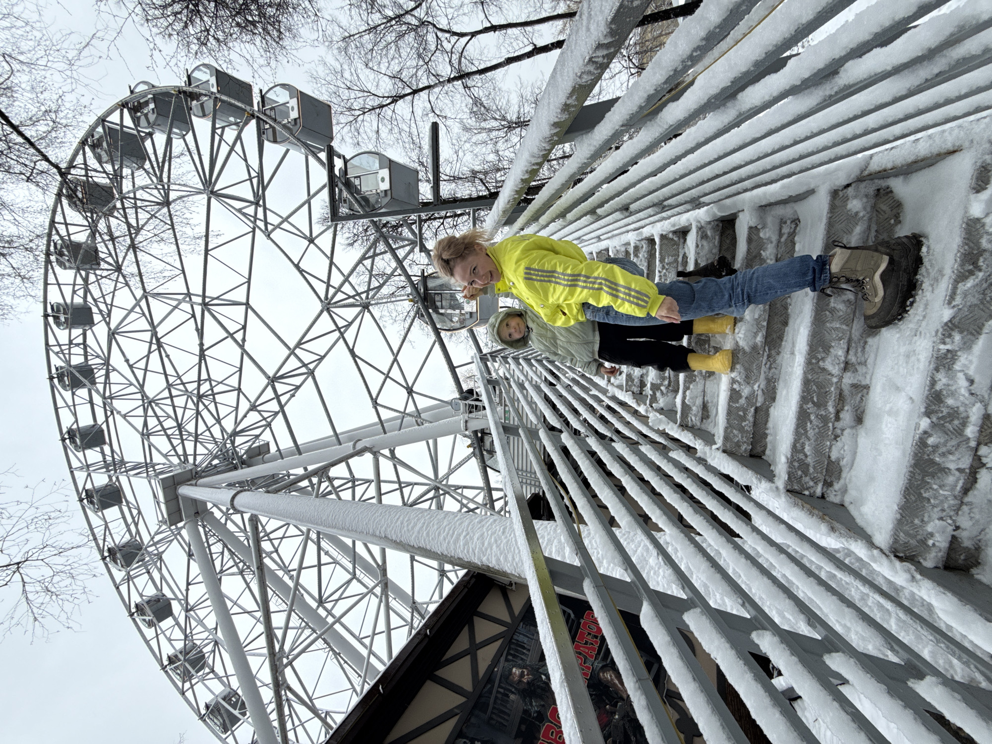 The image shows two individuals, likely a mother and child, positioned on a snowy metal staircase with a Ferris wheel in the background. The staircase railing is covered in snow. The mother is wearing a bright yellow jacket, blue jeans, and dark boots, looking at the camera and gesturing with her hand. The child, standing behind her, is wearing a light-colored jacket, black pants, and yellow boots.
The Ferris wheel is a large, gray metal structure with passenger cars visible. Bare trees are noticeable in the background. A building with signage is partially visible at the bottom of the frame.
Based on the presence of the Ferris wheel and the architectural style of the nearby buildings, the location appears to be in the city of Perm, Russia, within or near the Gorky Park. The scene suggests a winter setting due to the snow coverage.